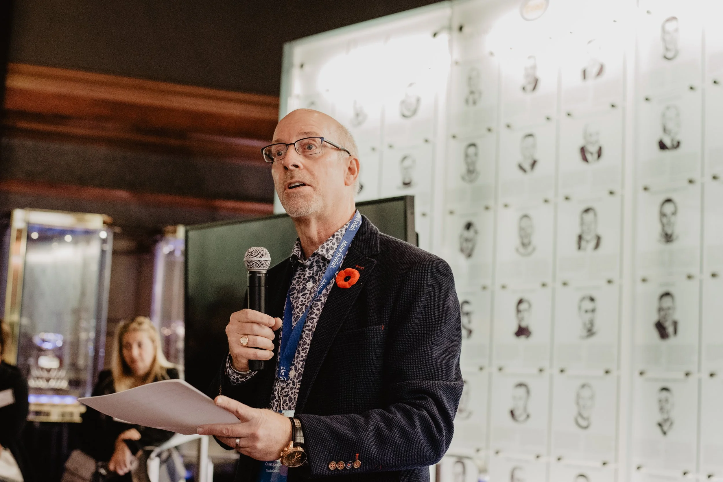 A man with glasses speaking into a microphone while holding papers, standing in front of a wall with black-and-white portrait sketches, with a woman in the background.