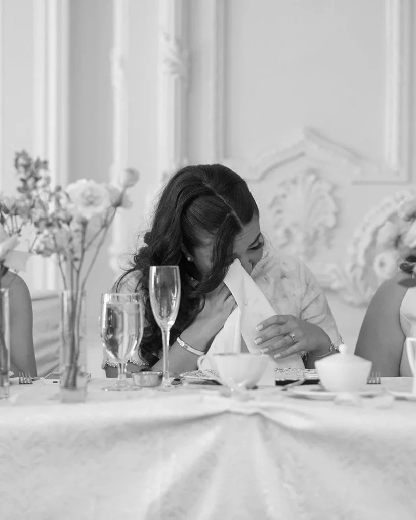 A woman with dark, wavy hair seated at a decorated table, holding a napkin to her face, with a smile, surrounded by champagne glasses, cups, and floral arrangements in a bright, elegant room.