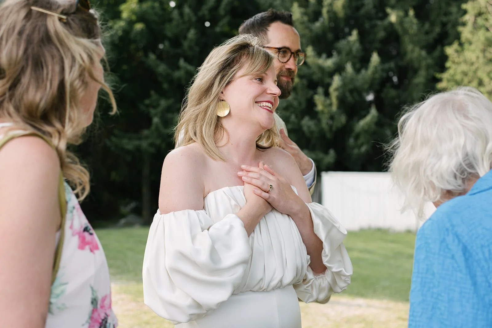 A woman in an off-shoulder white dress with blonde hair and earrings looks happy and emotional, holding her hands to her chest, surrounded by other women outdoors.