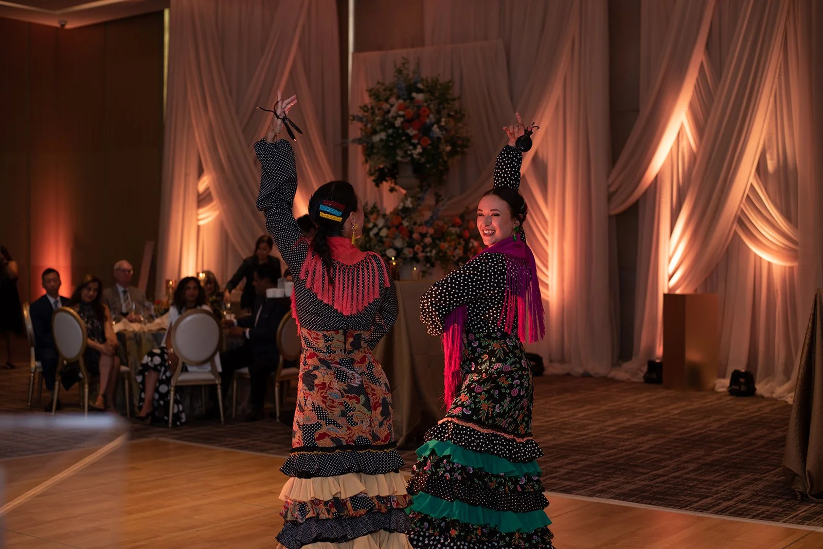 Two women in vibrant, colorful traditional dresses dancing at a formal event, with an audience seated at tables in the background and elegant drapery and floral arrangements on the wall.