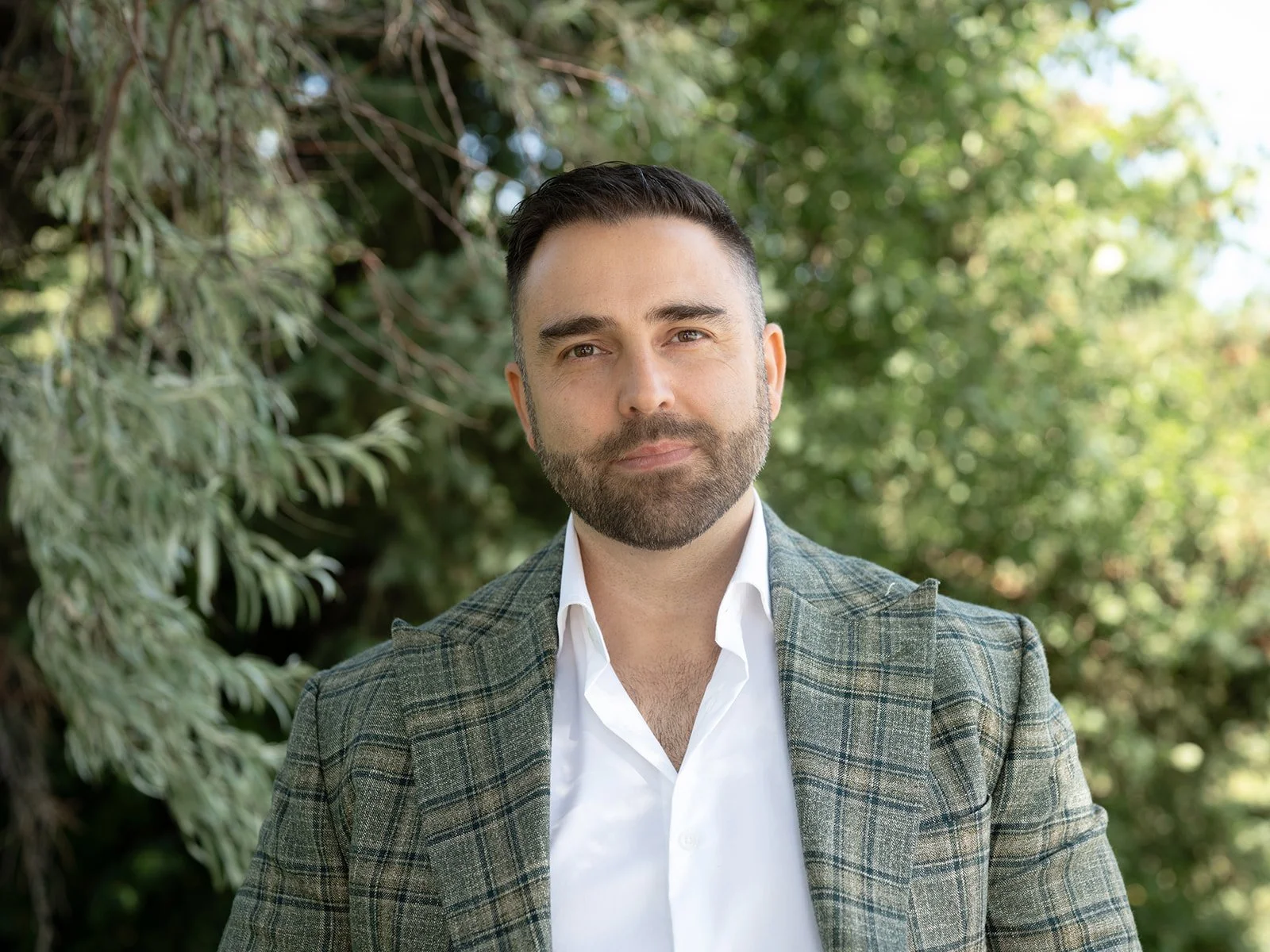 An outdoor corporate headshot of a man with dark hair and a beard, wearing a white shirt and a plaid blazer, standing outdoors with green foliage in the background.