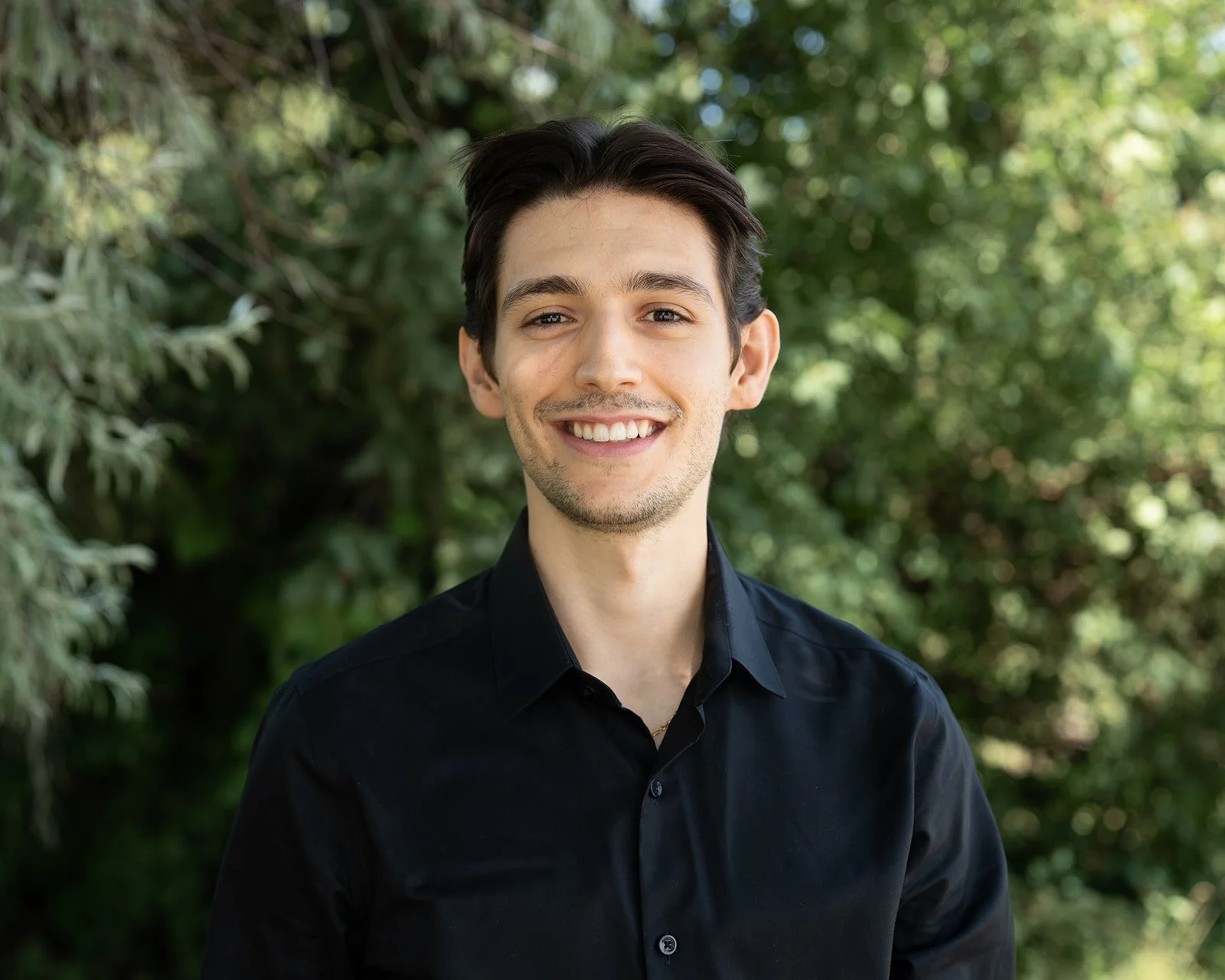 A corporate headshot of a young man with dark hair and a light beard, smiling at the camera, wearing a black shirt, outdoors with green trees in the background.