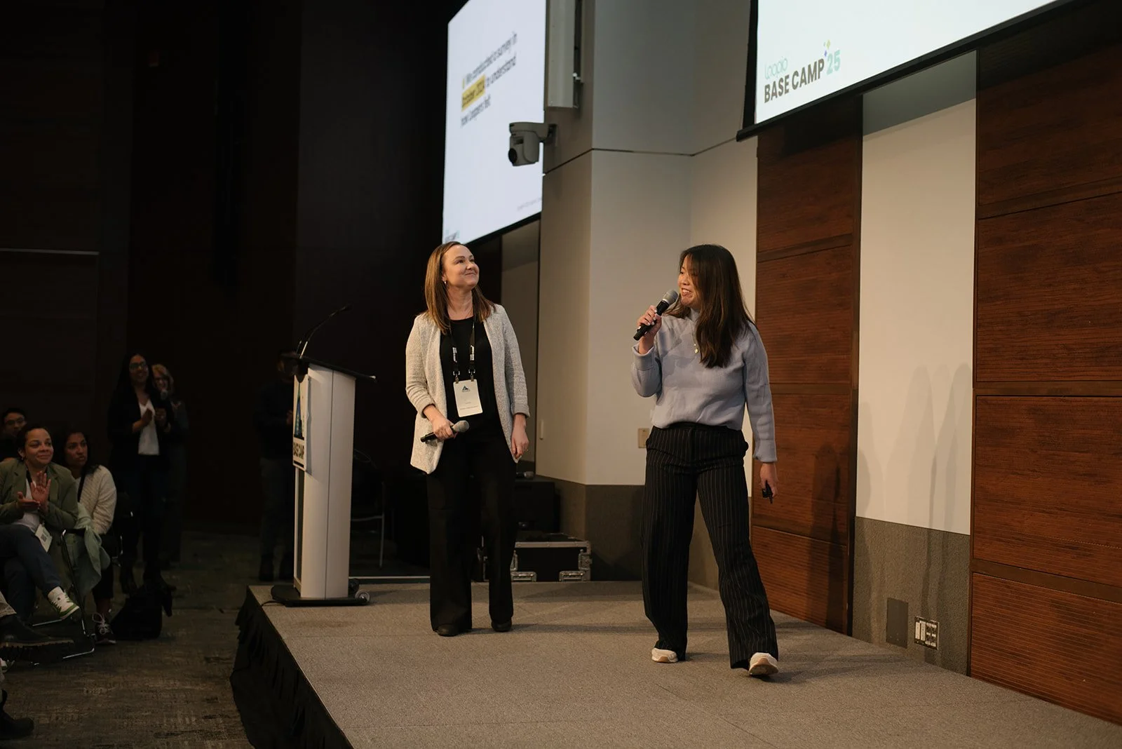 Two women speaking on stage at a conference, with one holding a microphone. An audience watches them, and a large screen displays a logo with the words "Base Camp."