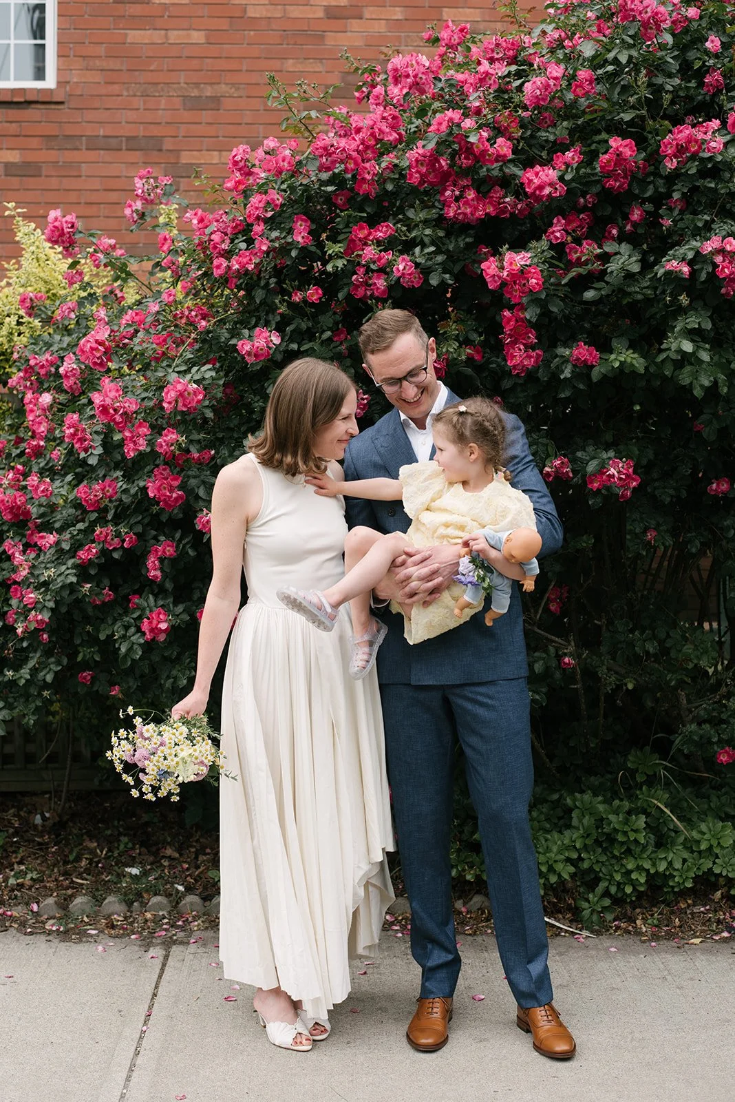 A family of four standing outdoors in front of a large blooming pink bush, smiling and interacting. The woman is holding a bouquet of flowers, and the man is holding their young daughter in a yellow dress and a doll.