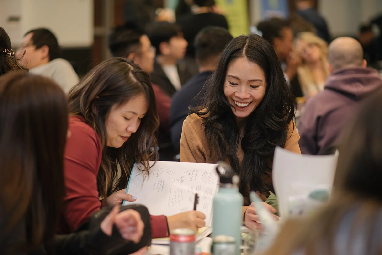 A group of people sitting at a table during a meeting or workshop. One woman is writing in a notebook while another woman next to her is smiling and looking at her.