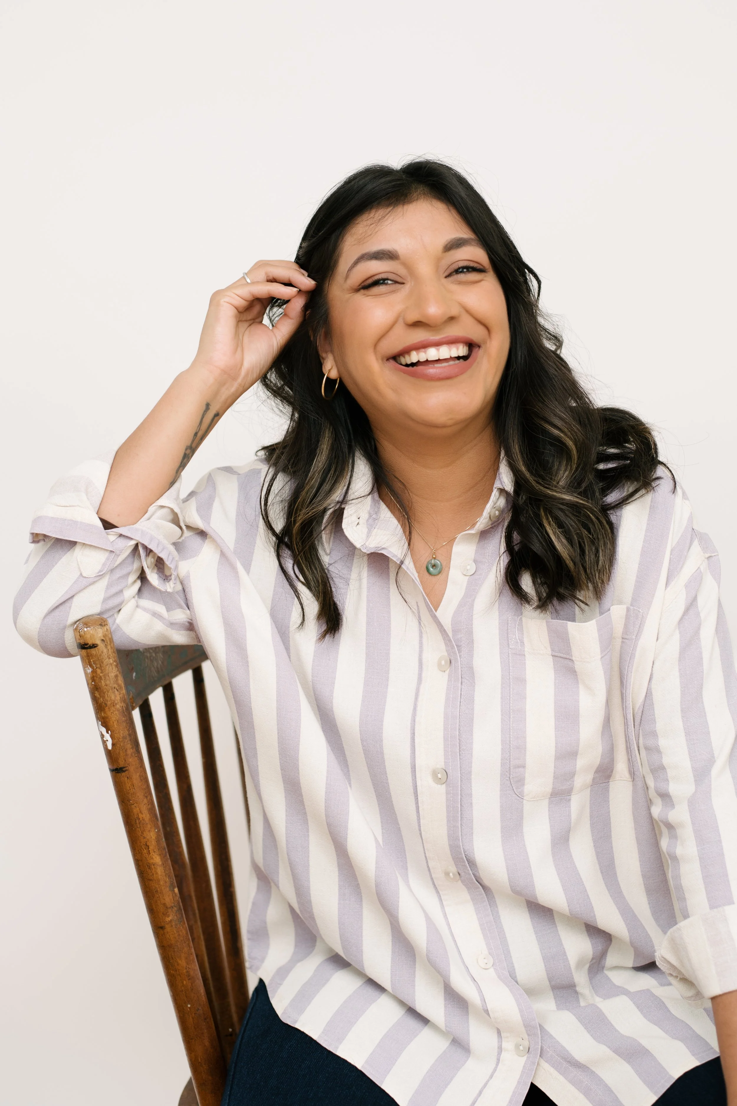 A woman with long dark hair, wearing a striped button-up shirt, sitting on a wooden chair, and smiling at the camera.