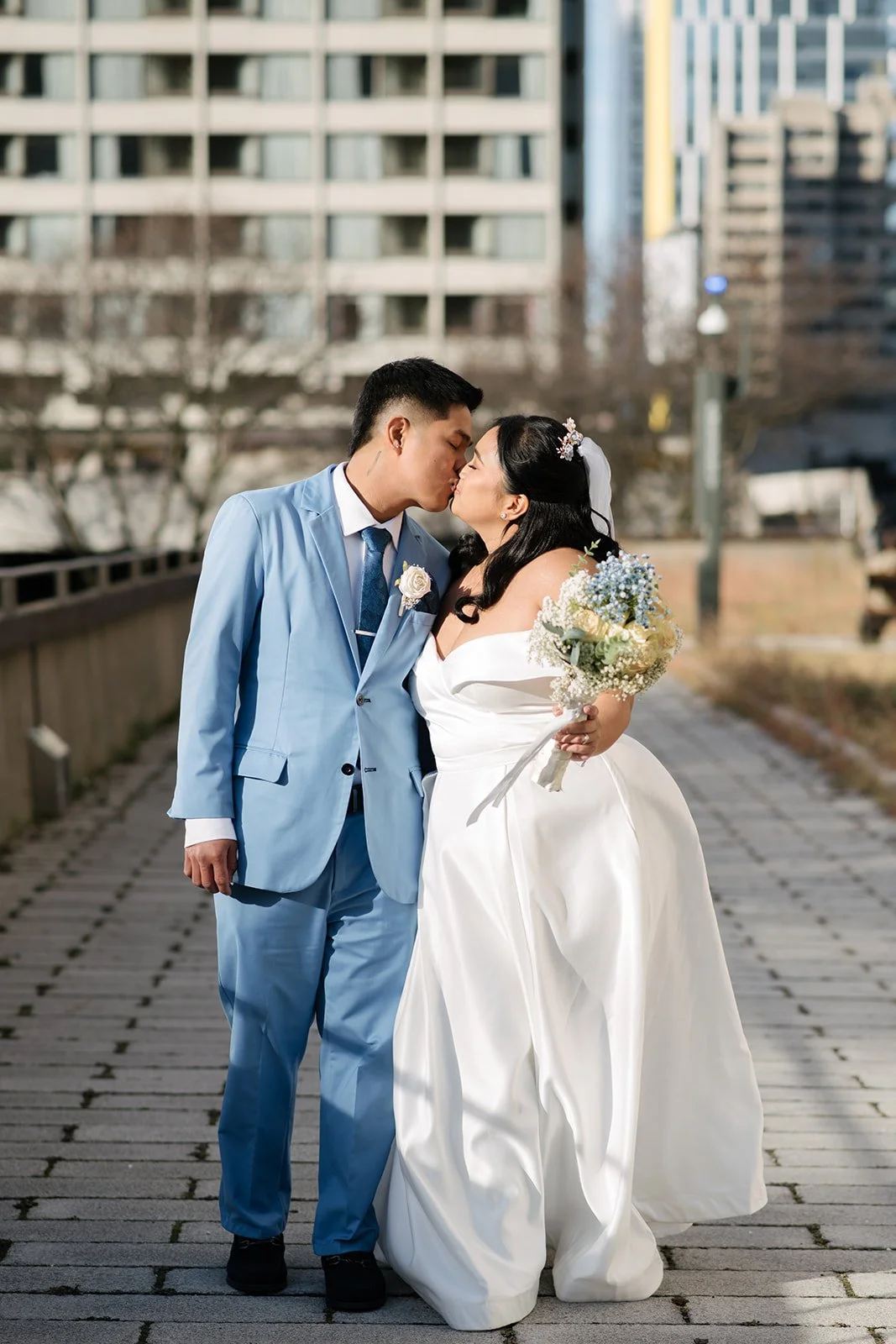 A bride and groom sharing a kiss outdoors at Toronto City Hall.  The groom is wearing a light blue suit and the bride is wearing a long white wedding dress.