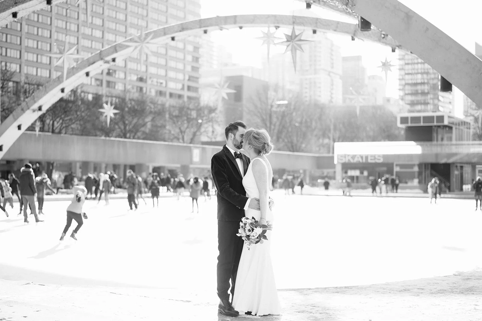 A black and white photo of a couple in wedding attire sharing a kiss on an outdoor ice skating rink, with city buildings and skaters in the background.