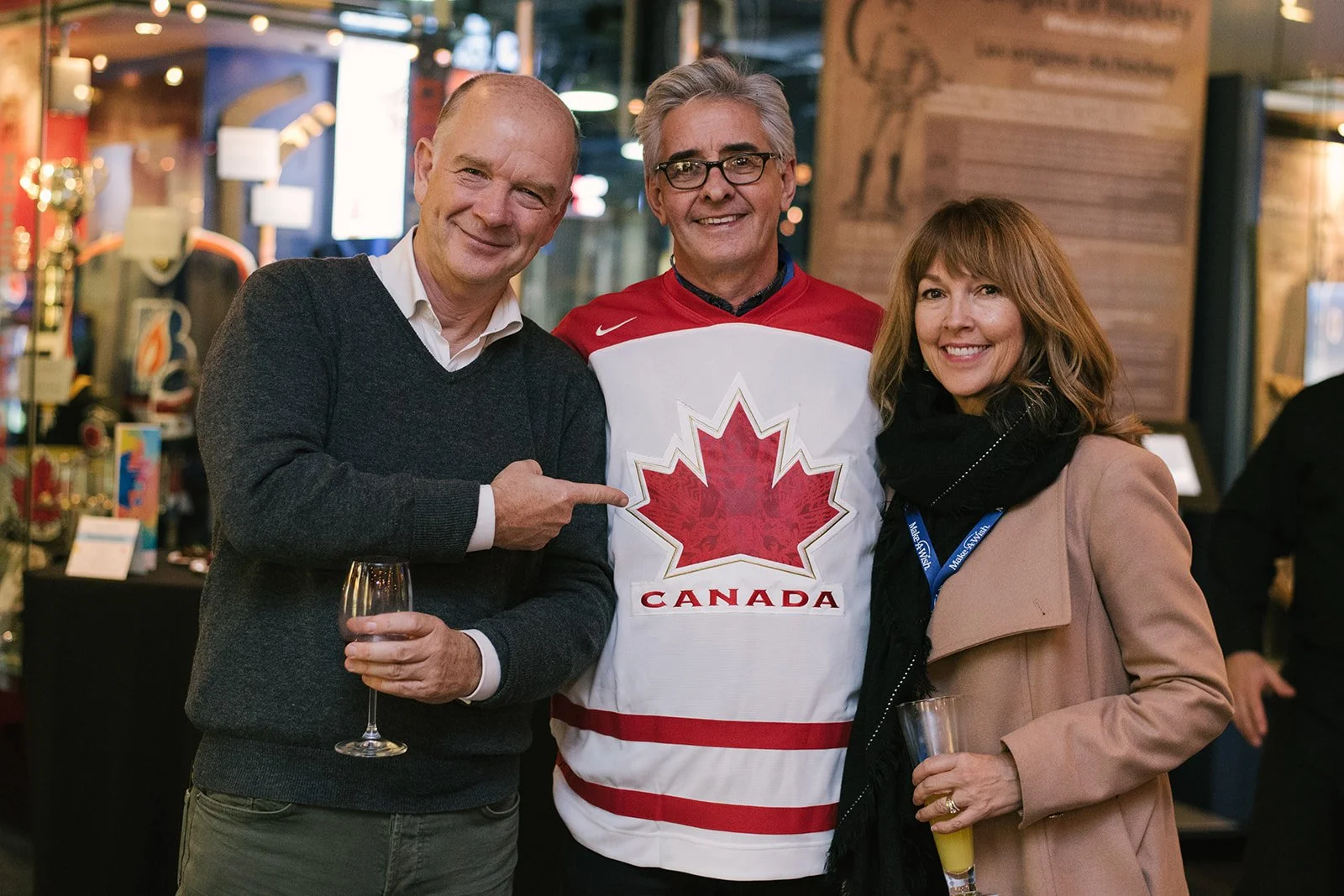 Three people smiling at a social event, with the man in the middle wearing a Canadian sports jersey with a red maple leaf and the word 'Canada'. The man on the left is holding a glass of wine, and the woman on the right is holding a drink. They are indoors with warm lighting and blurred background.