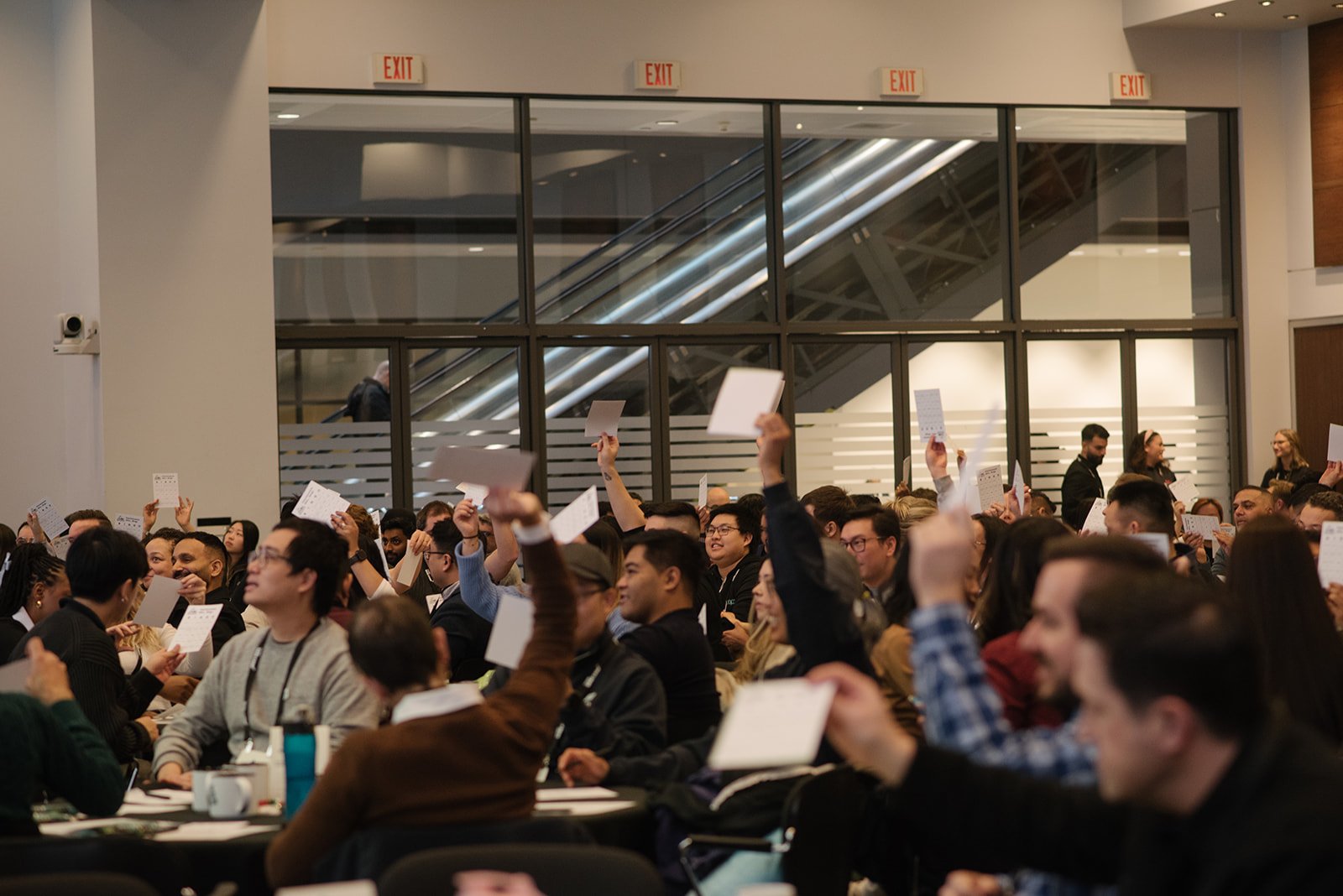 Large conference room filled with attendees raising their hand to ask questions or participate, with papers and laptops on tables.