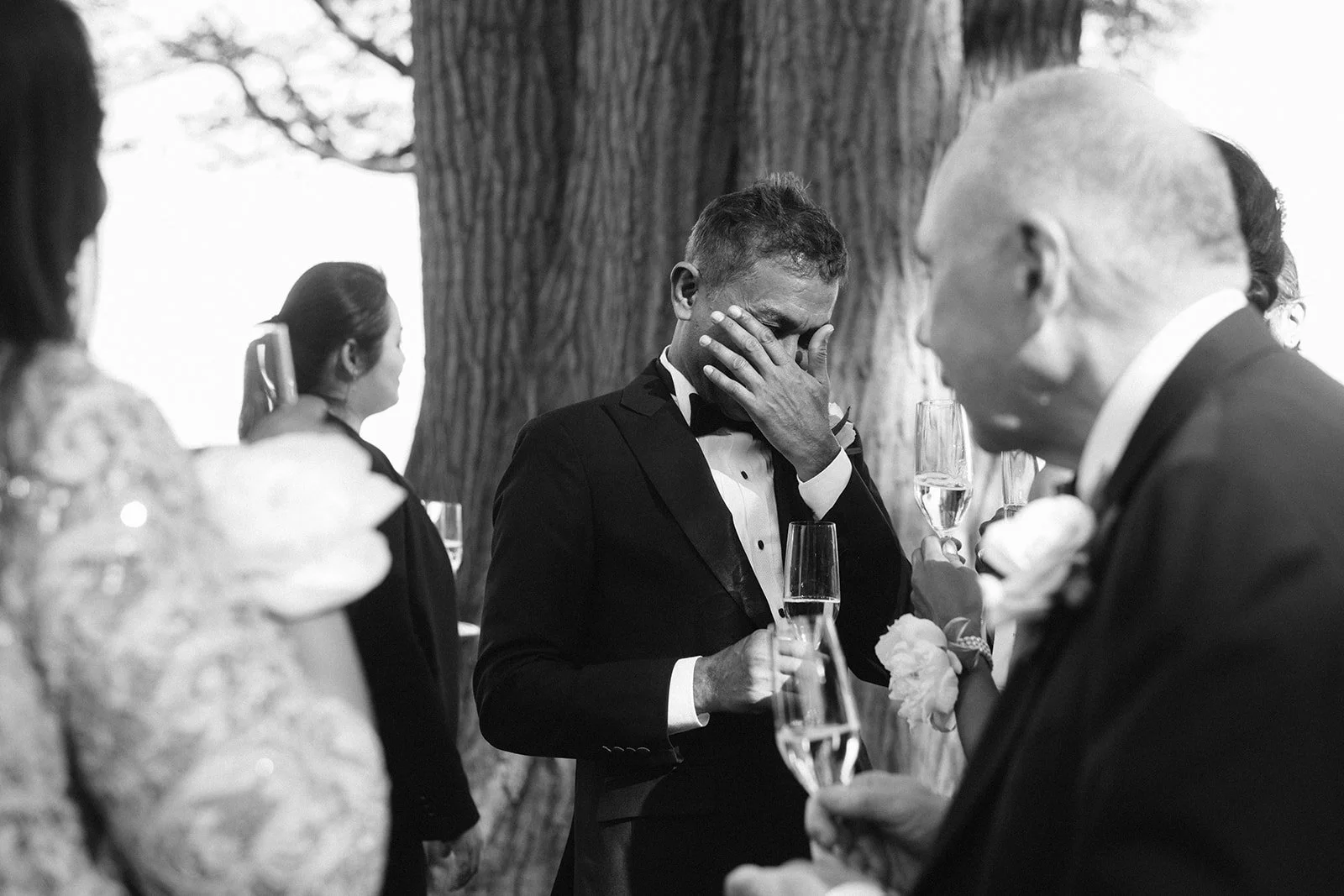 A black and white photo of a group of people at a celebration, with one man dressed in a tuxedo wiping away tears, holding a glass of champagne, surrounded by others holding champagne glasses, in front of a large tree.