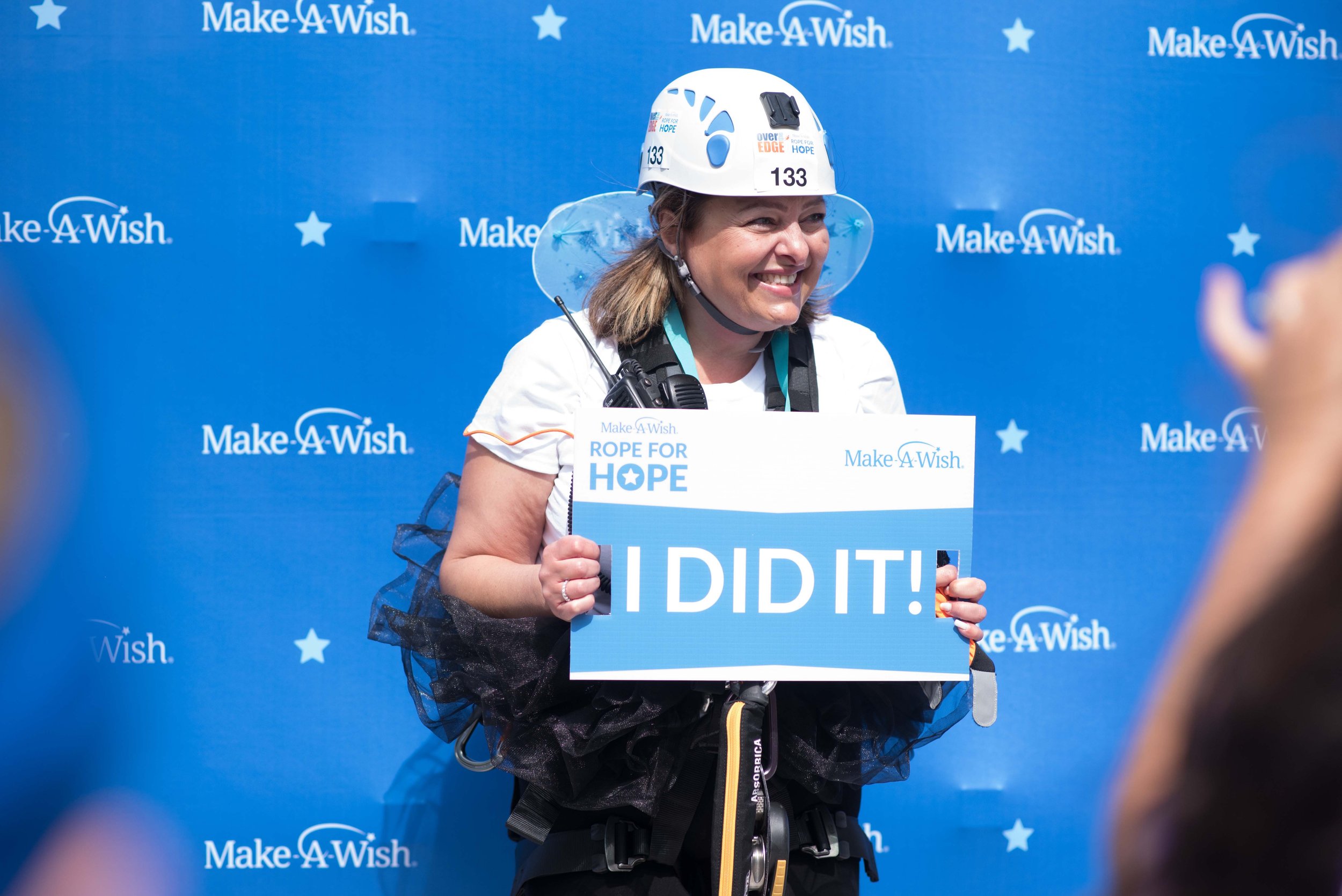 Woman smiling holding a sign that says "I DID IT!" at a Make-A-Wish event, wearing a climbing helmet and harness, standing in front of a blue backdrop with the Make-A-Wish logo.