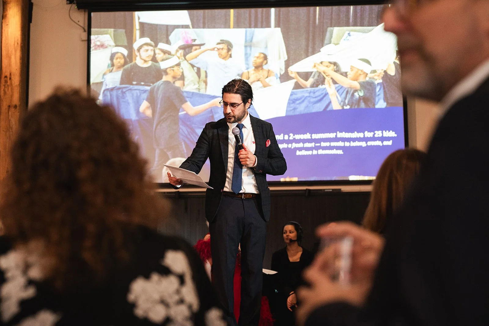 A man in a suit and glasses is holding a microphone and reading from a paper while standing in front of a large screen showing a photo of children wearing hats and holding a banner. Several people are seated in the foreground, listening, and among them is a woman wearing a headset.