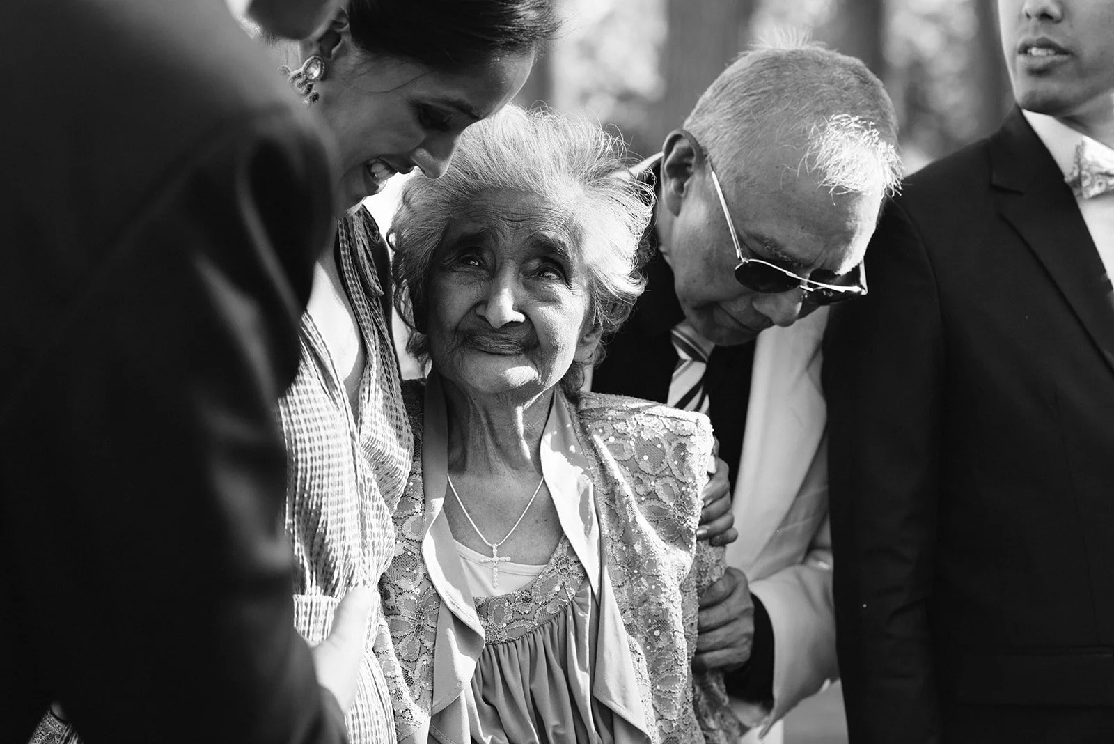 Black-and-white photo of four people in formal attire, mainly focusing on an elderly woman looking up at a woman and man beside her. The woman, with gray hair and an ornate outfit, appears emotional. The others are leaning toward her, sharing a momen