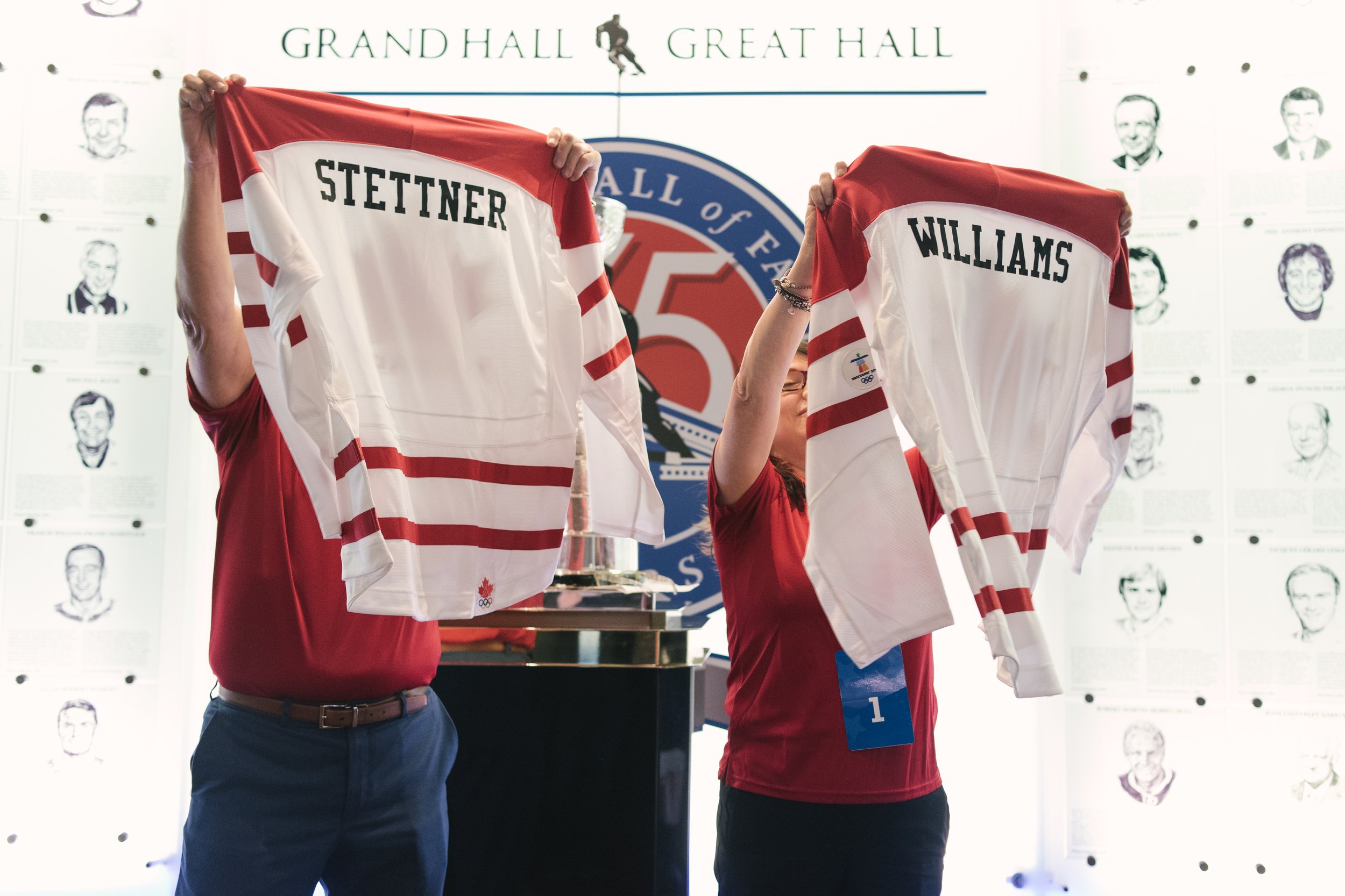 Two people holding up hockey jerseys with names 'Stettner' and 'Williams' at the Hall of Fame, with portraits and the 'Great Hall' sign in the background.