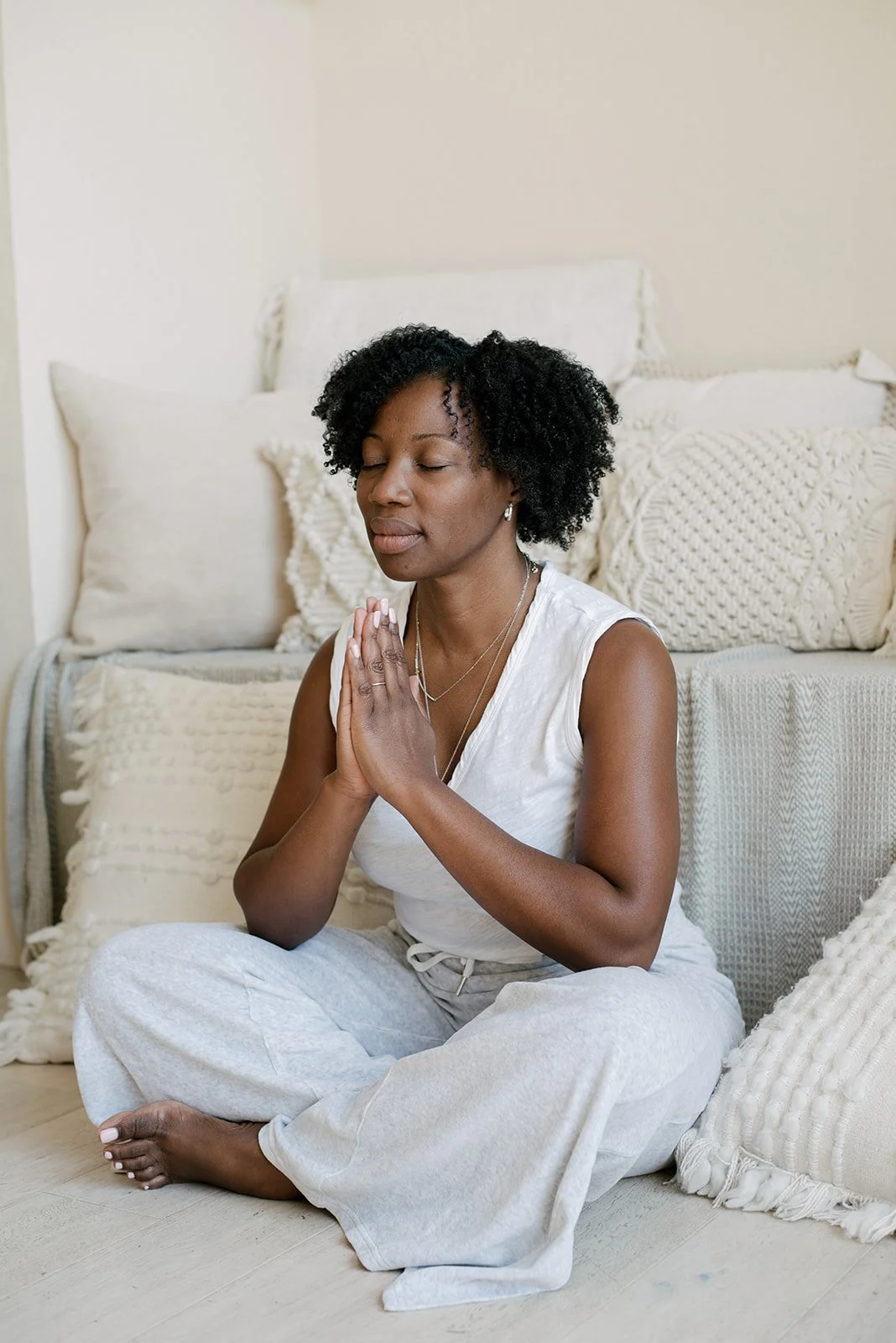 A woman practicing meditation or prayer, sitting cross-legged on the floor with her eyes closed and hands pressed together in prayer, surrounded by stuffed pillows in a cozy, light-colored room in Mint Room Studios in Toronto.