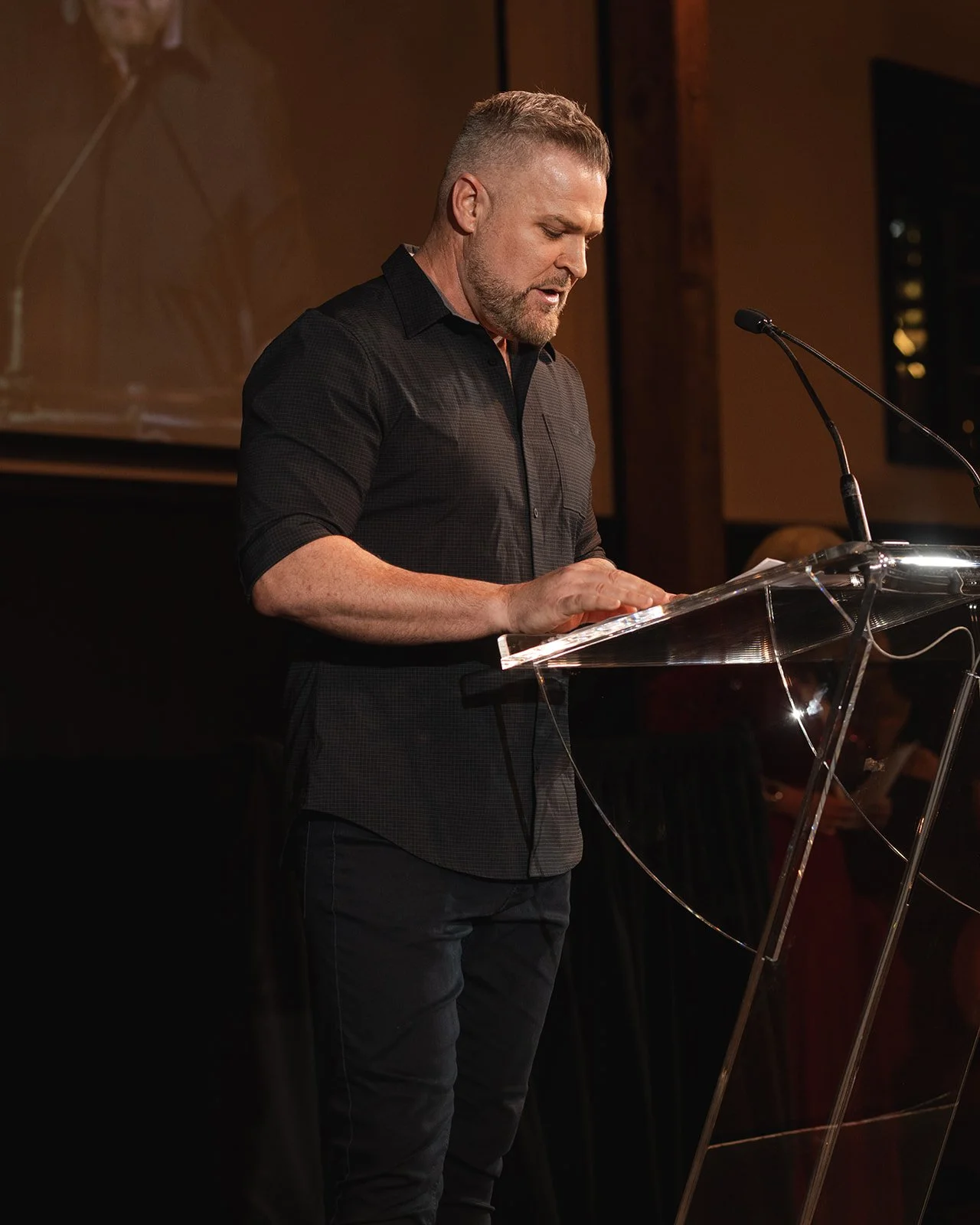 A man with short hair and beard wearing a black shirt standing at a transparent podium, speaking into a microphone, at a gala in Toronto.