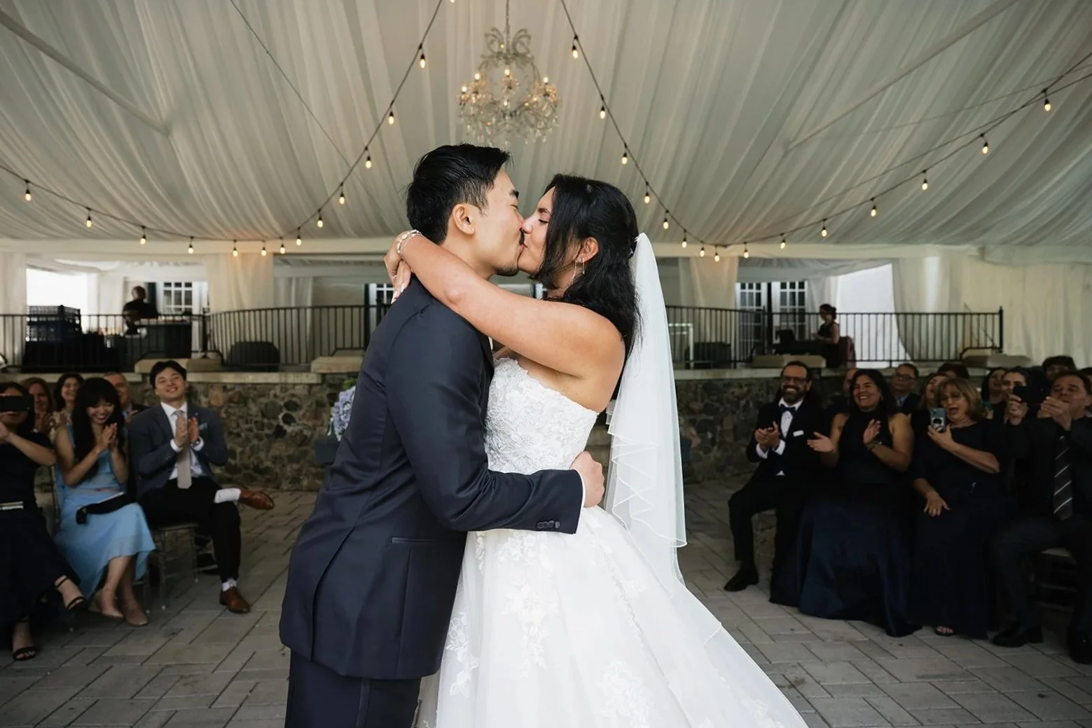 A newlywed couple is kissing during their wedding ceremony, surrounded by friends and family at the Guild Inn wedding venue with string lights and a chandelier overhead.