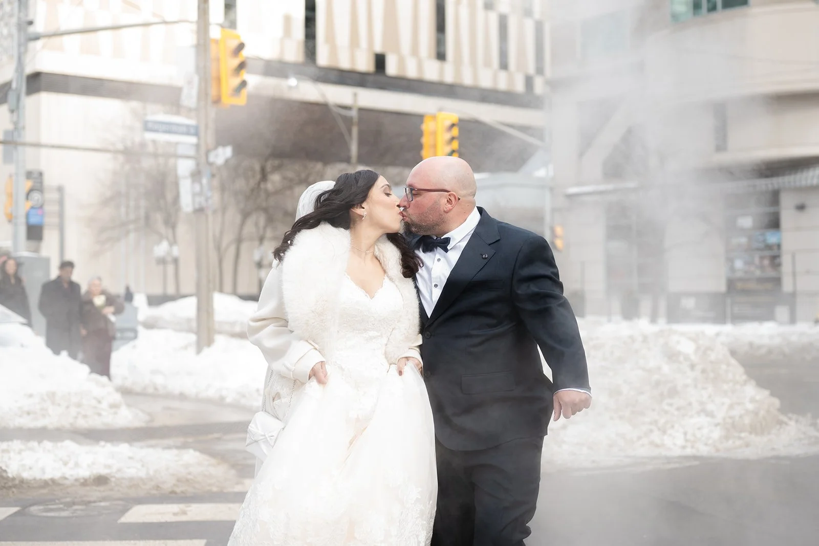 A bride in a white wedding dress and a groom in a black tuxedo sharing a kiss outdoors in a snowy city street on their way to get married at Toronto City Hall