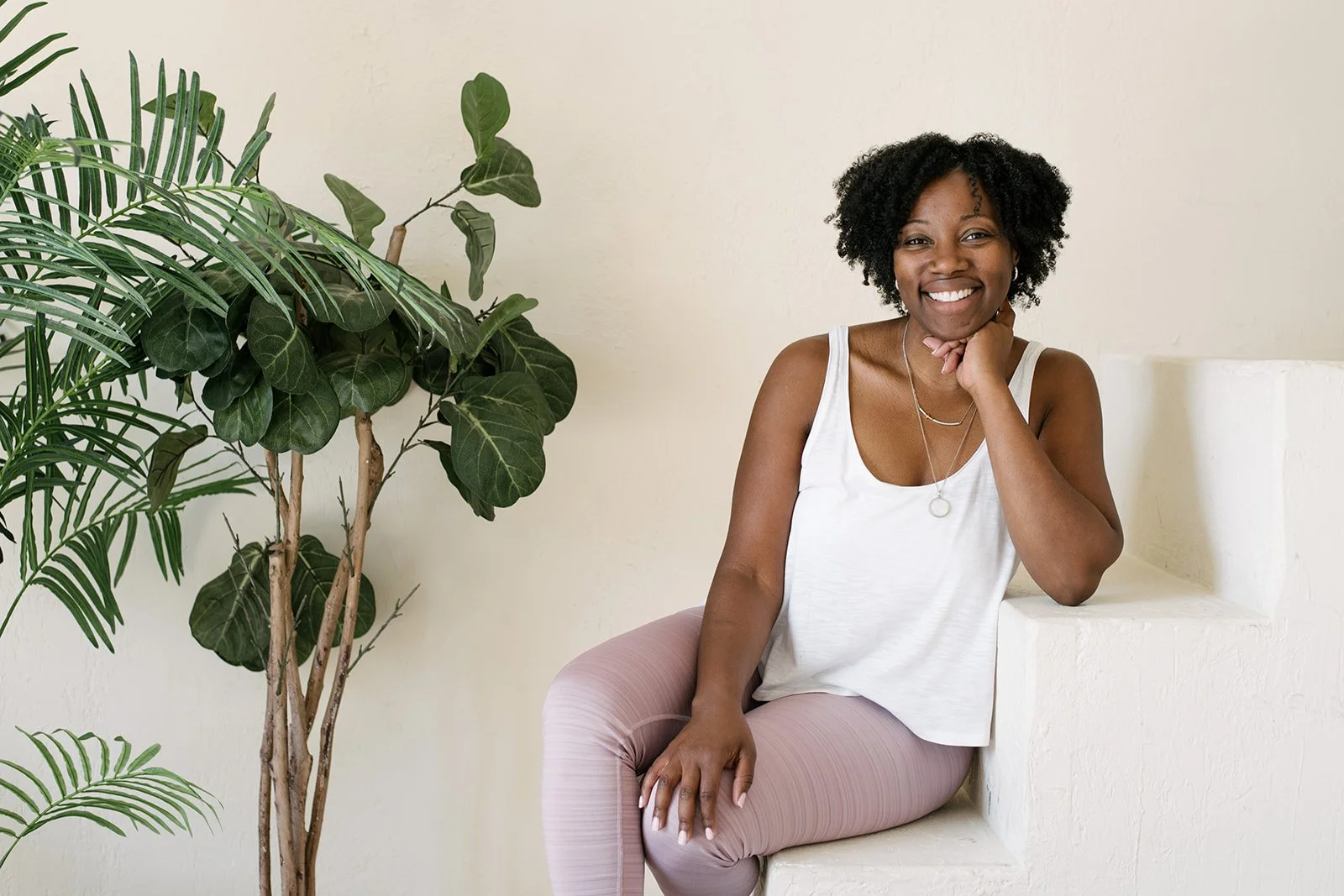 A smiling woman with curly black hair sitting on a white built-in bench near a large green plant at Mint Room Studios in Toronto.