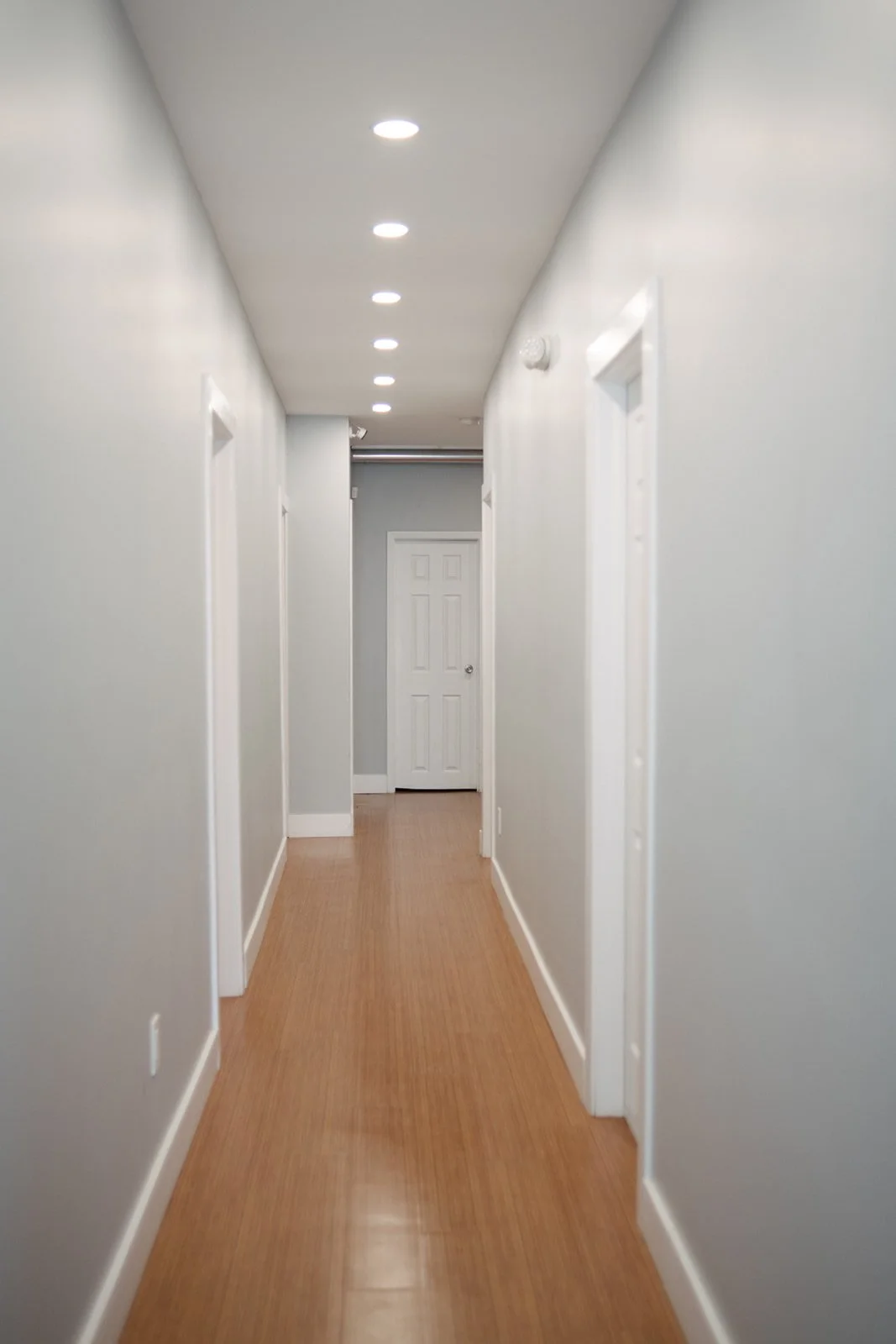 A hallway with white walls, wood flooring, and white doors, illuminated by recessed ceiling lights.