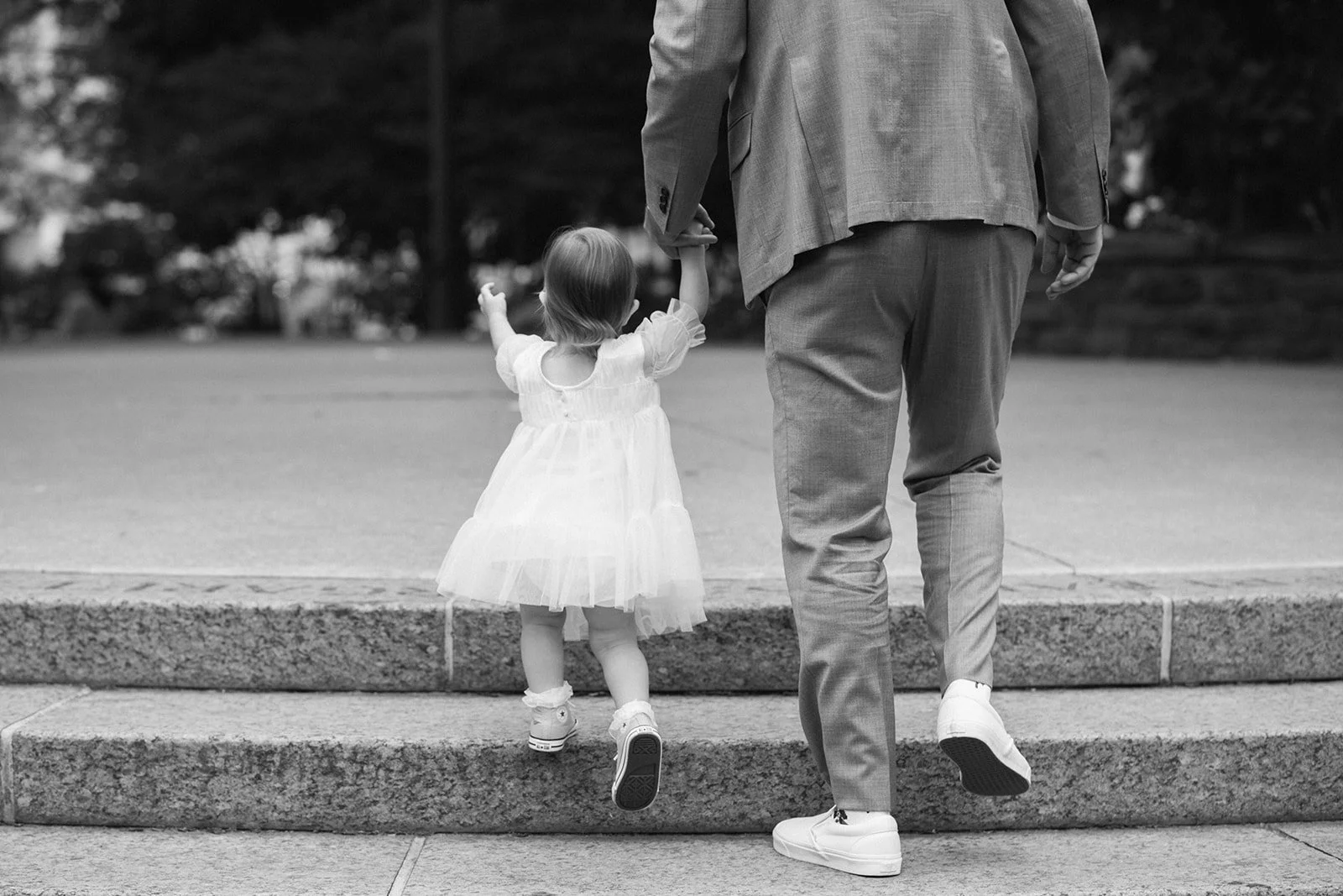 A young girl in a white dress and sneakers is holding hands with her father, as they walk up some steps outdoors at Toronto Music Garden before a wedding.