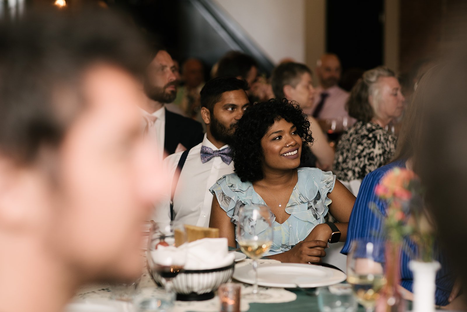 A group of people sitting at a table, attending a wedding reception at The Broadview Hotel, with some people smiling and paying attention to speeches off-camera.
