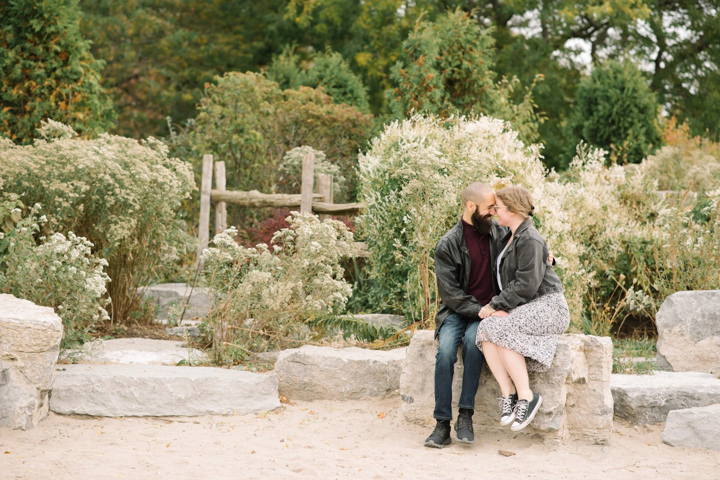 A couple sitting on a stone bench in a garden surrounded by white flowering bushes, with trees in the background. They are close, touching foreheads and smiling at Sunnyside Beach in Toronto.