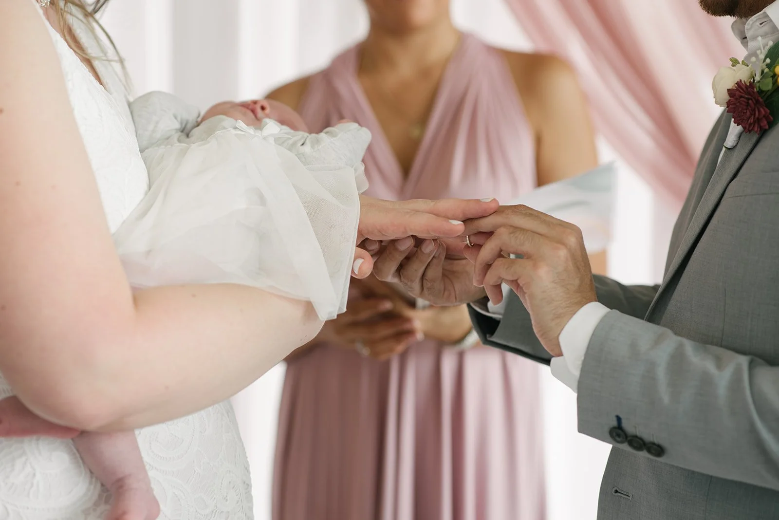 A wedding ceremony where a bride is holding a sleeping baby, and a groom is placing a wedding ring on her finger with an officiant and bridesmaid present.