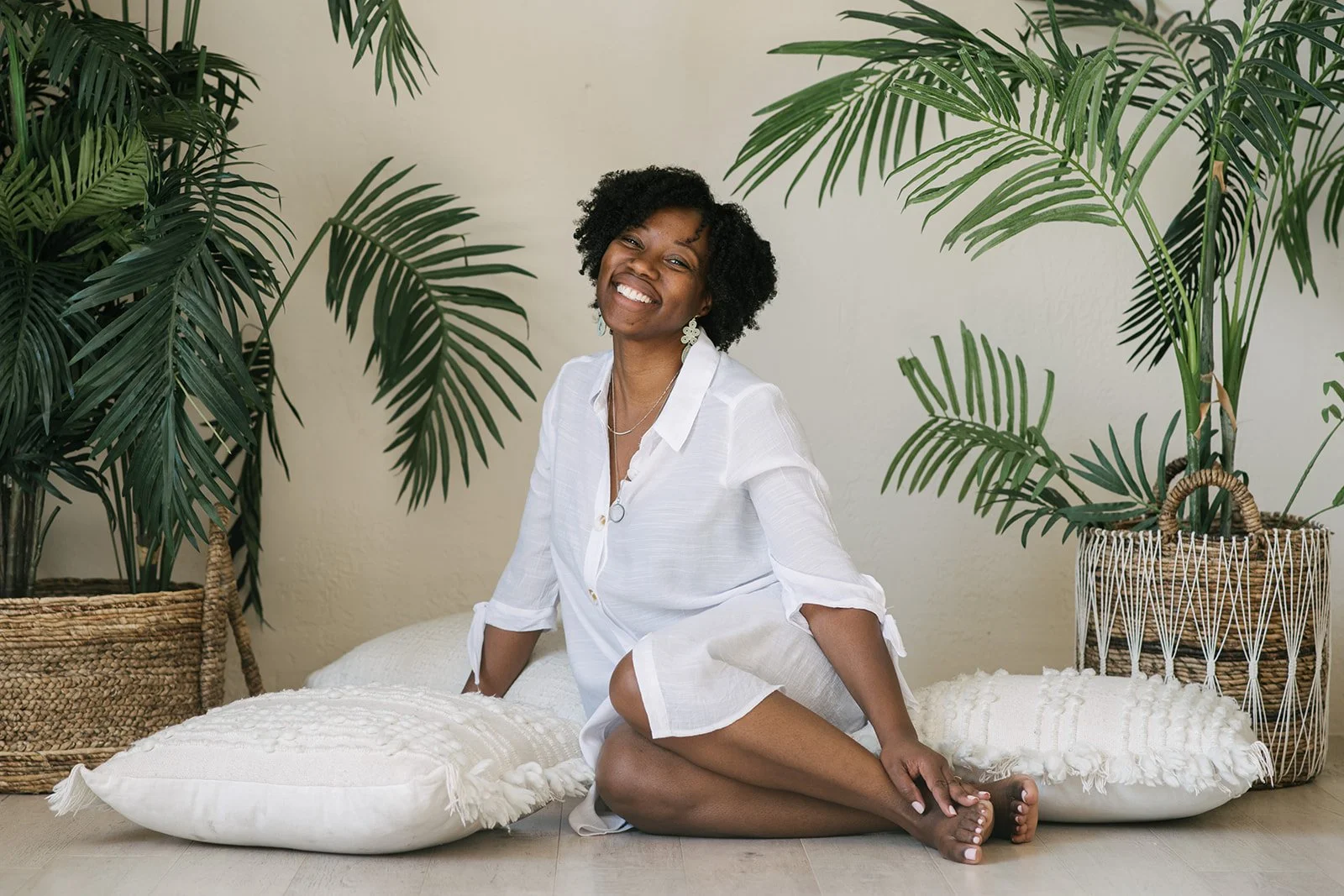 A woman with short curly black hair, wearing a white shirt, sitting on light-colored pillows on the floor between large potted plants with tall green leaves, smiling at the camera in Mintroom Studios in Toronto