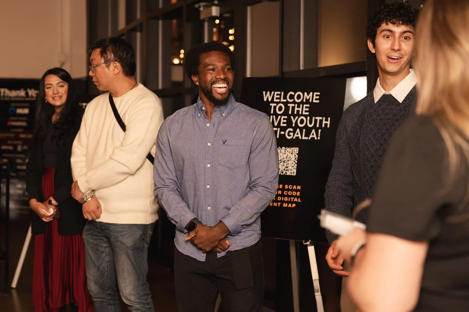 Group of people standing the Thrive Youth Anti-Gala, engaging in conversation, with a poster board displaying QR code and event details behind them.