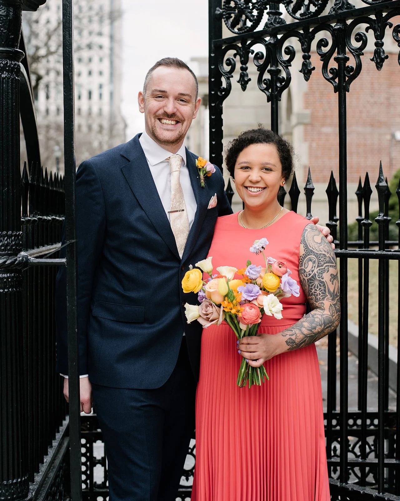 A smiling couple standing together outdoors near an ornate black iron gate, with the woman holding a colorful bouquet of flowers, both dressed in formal attire, during daytime.