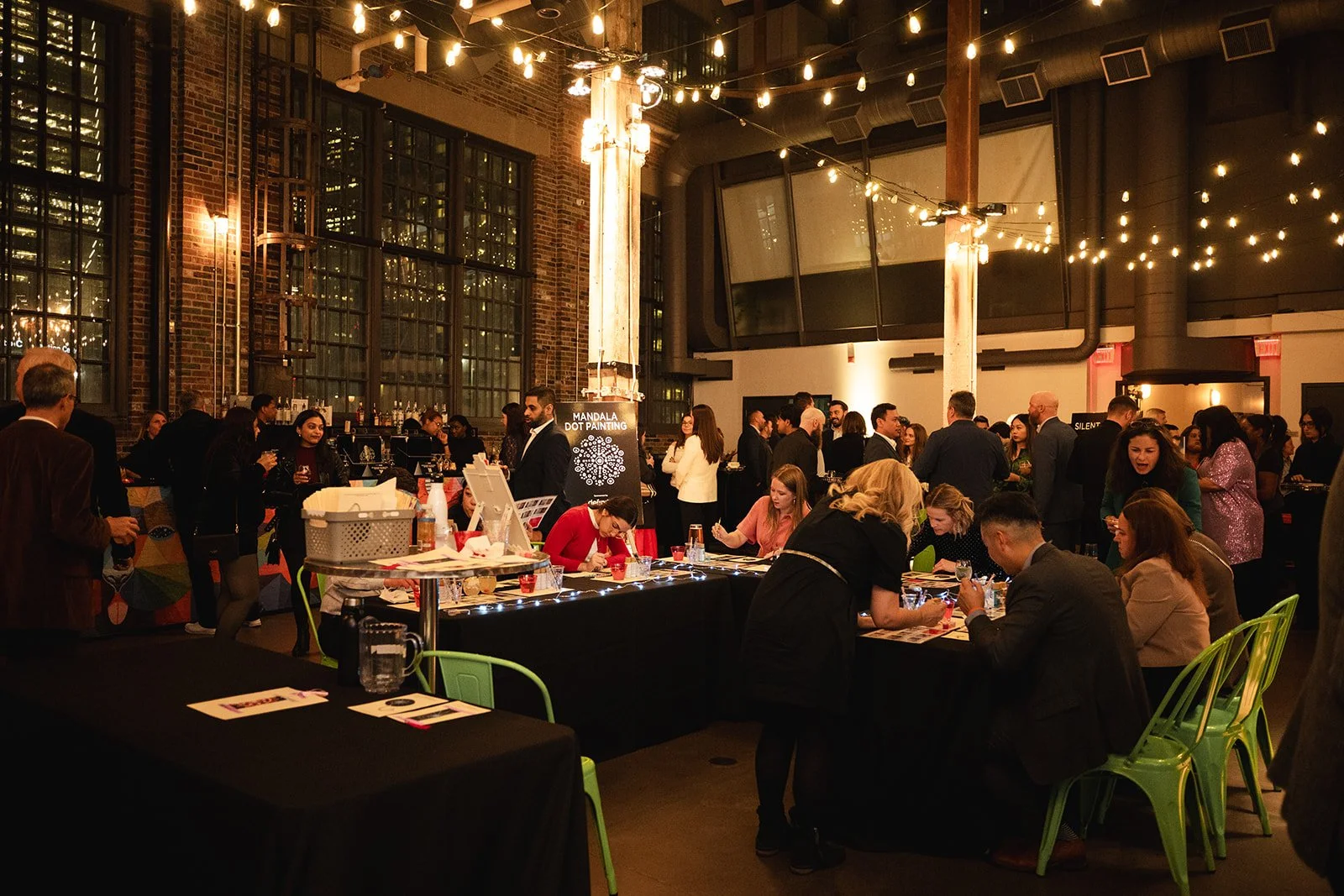 Indoor event with people socializing, tables with arts and crafts, large windows, exposed brick walls, string lights, and a display sign for a mandala dot painting activity.