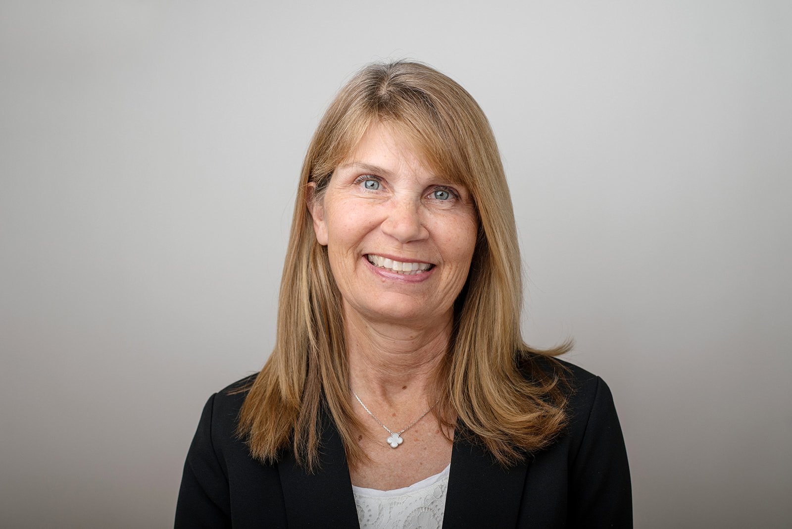 Smiling woman wearing a black blazer and white top, in front of a grey background