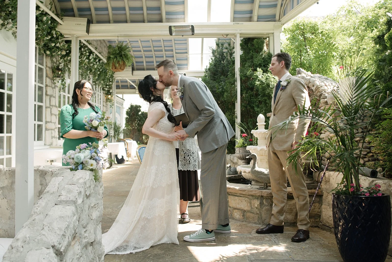 A wedding ceremony featuring a couple kissing, surrounded by an officiant and wedding party members, at Auberge du Pommier with greenery and a fountain.