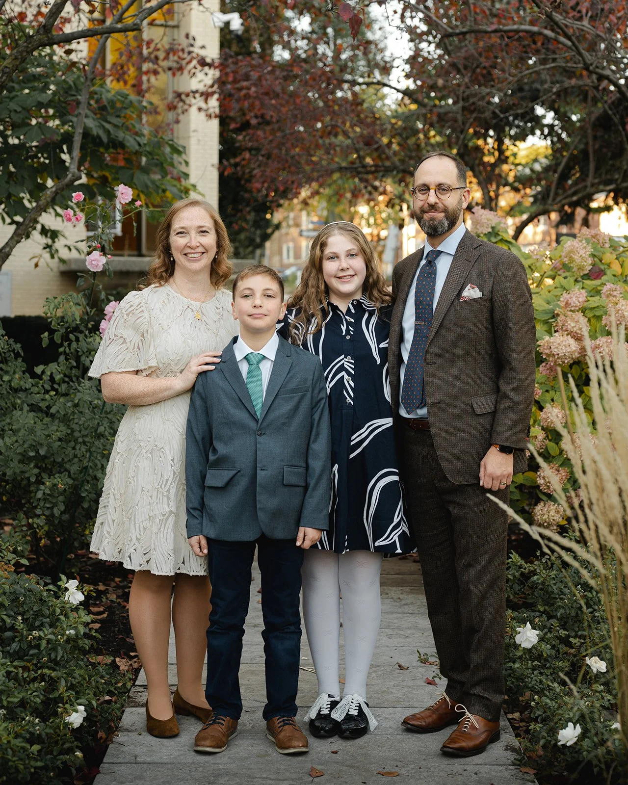 A family of four standing on the grounds of Beth Tzedec Congregation surrounded by trees and bushes with flowers, during fall, smiling for a photo.
