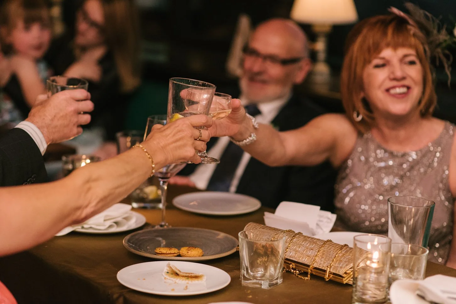 People celebrating with drinks at a dinner party, raising glasses in a toast. The scene includes a table with desserts, candles, and a group of smiling guests, some in formal attire.