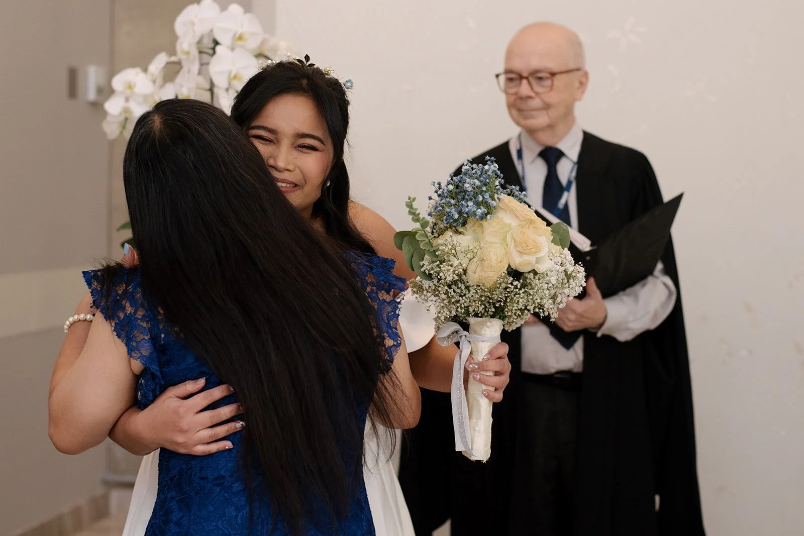 Two women hugging, one holding a bouquet of white and blue flowers, while a man in a judge's robe in the Toronto City Hall Wedding chambers stands in the background holding a black folder.