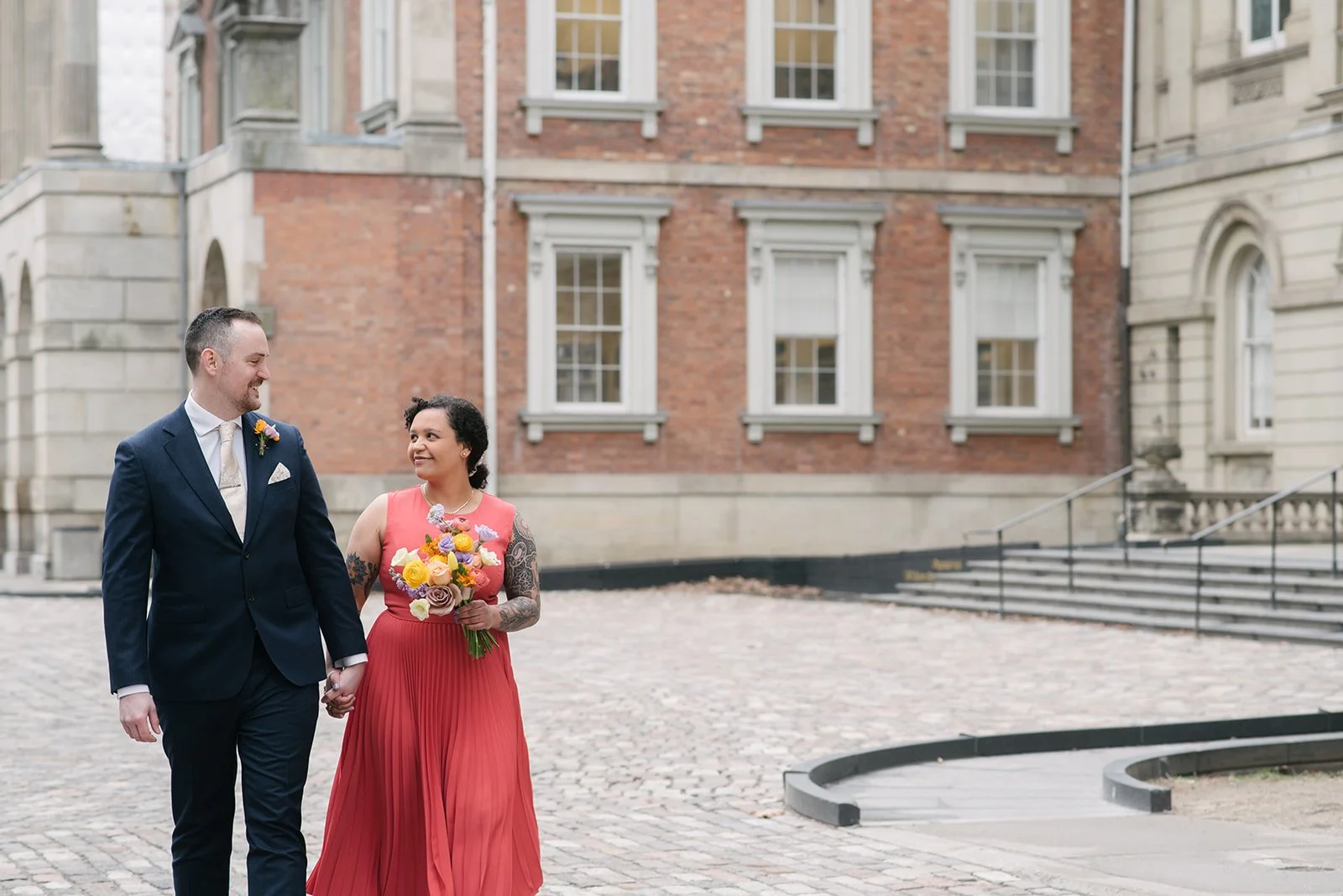 A man in a dark suit holding hands with a woman in a pink dress holding a colourful bouquet, walking together on a cobblestone pathway at Osgoode Hall near Toronto City Hall.