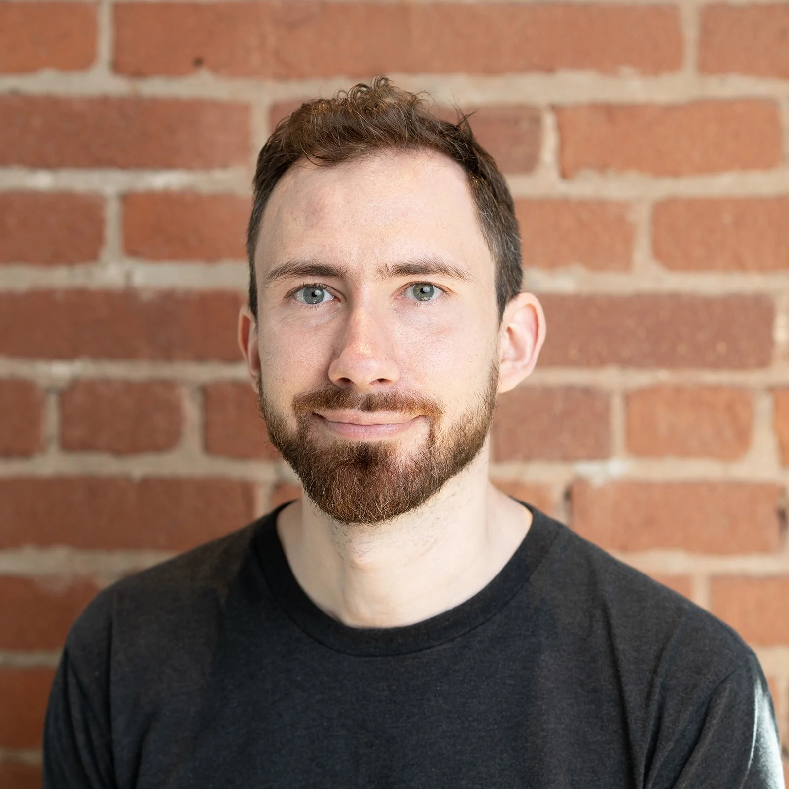 A corporate headshot of a man with light skin, blue eyes, a beard, and brown hair, standing against a red brick wall. He is wearing a black T-shirt and smiling slightly.