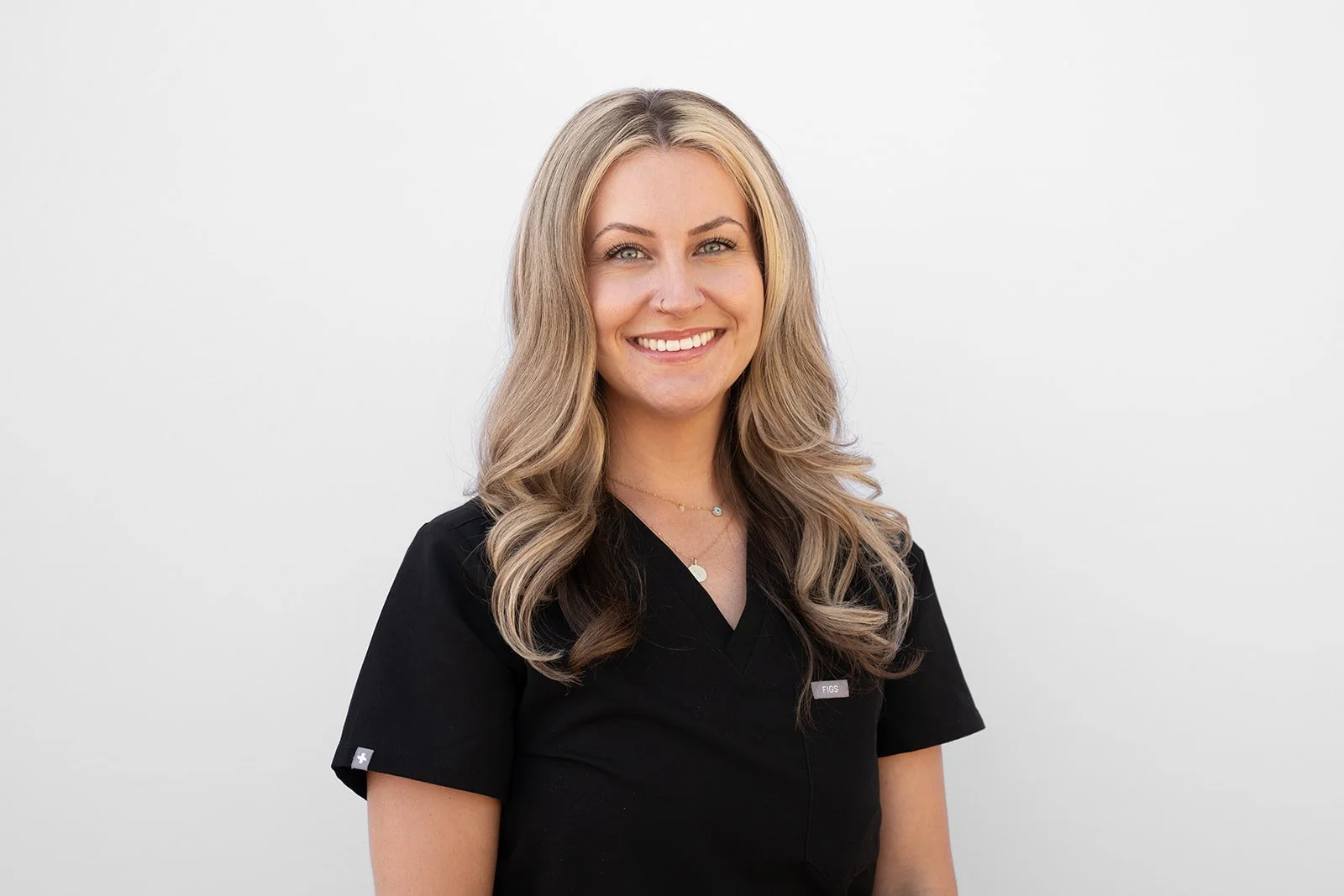 A headshot of a young woman with long, blonde, curly hair, smiling, wearing a black medical uniform against a plain white background.