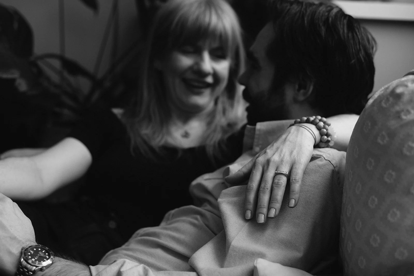 A woman with light-colored hair smiling and leaning over a man with dark hair, who is lying down. The woman's hand, decorated with an engagement ring and beaded bracelet, rests on the man's shoulder.