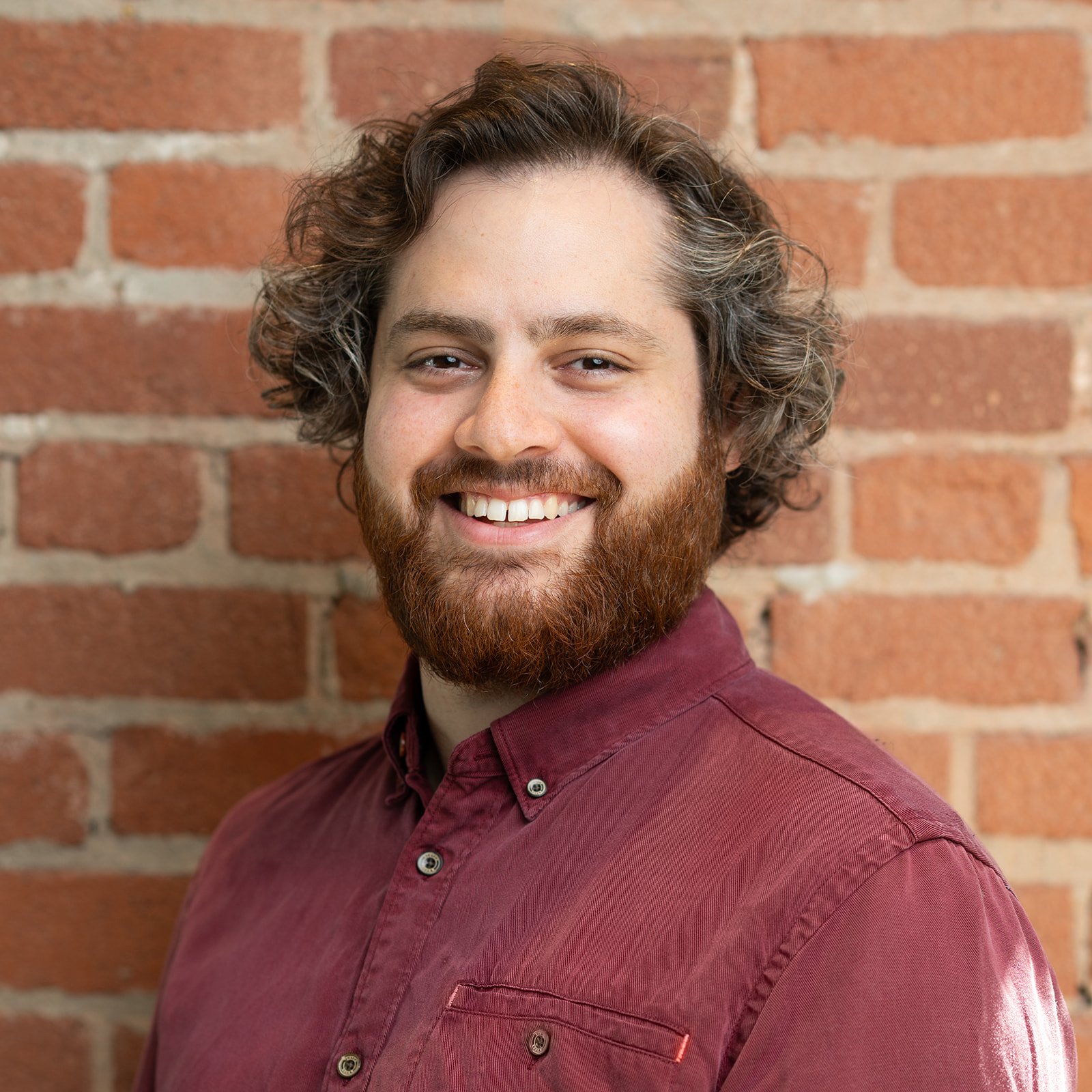 A corporate headshot of a smiling man with curly hair and a beard wearing a maroon button-up shirt, standing in front of a brick wall.
