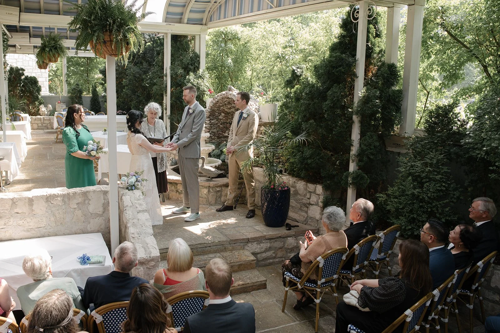 A wedding ceremony featuring a couple kissing, surrounded by an officiant and wedding party members, at Auberge du Pommier with greenery and a fountain.