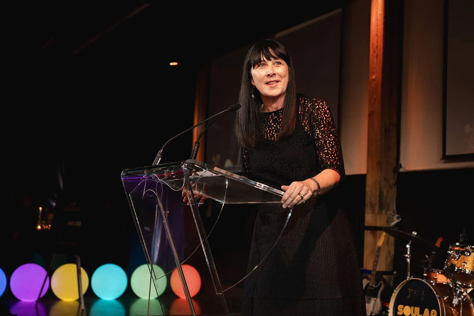 A woman with long dark hair, wearing a black lacy dress, stands at a clear glass podium speaking into a microphone during a Gala. There are colourful illuminated spherical lights on the floor behind her, and a drum set is visible in the background.