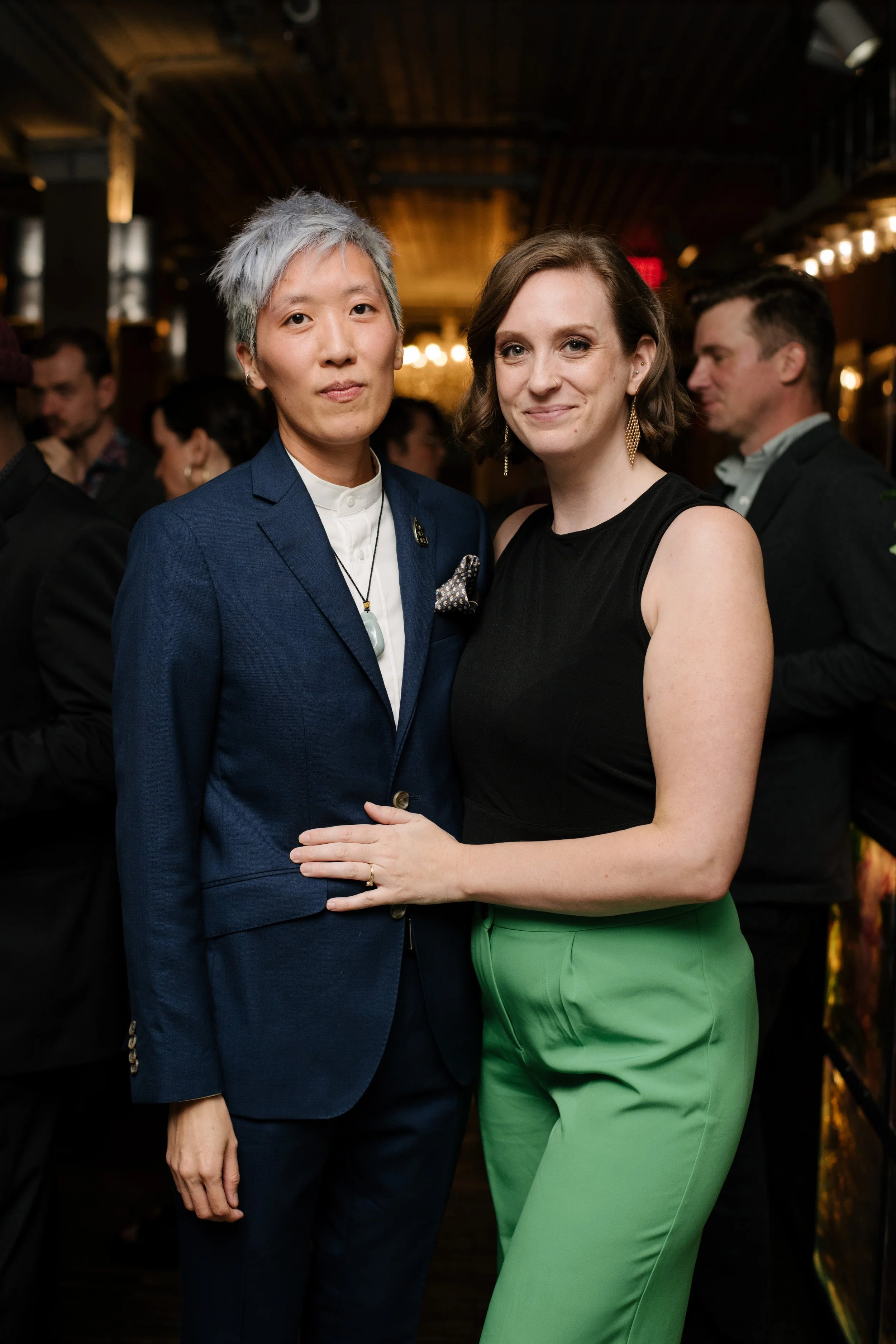 Two women posing together at an indoor event, one wearing a navy blazer and the other wearing a black sleeveless top and green pants, with other people in the background.