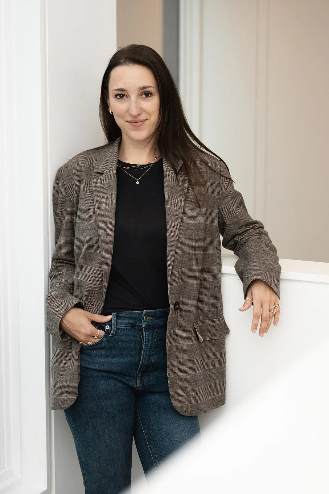 A woman with long dark hair, wearing a black top and a plaid blazer, stands against a white wall in a professional setting.