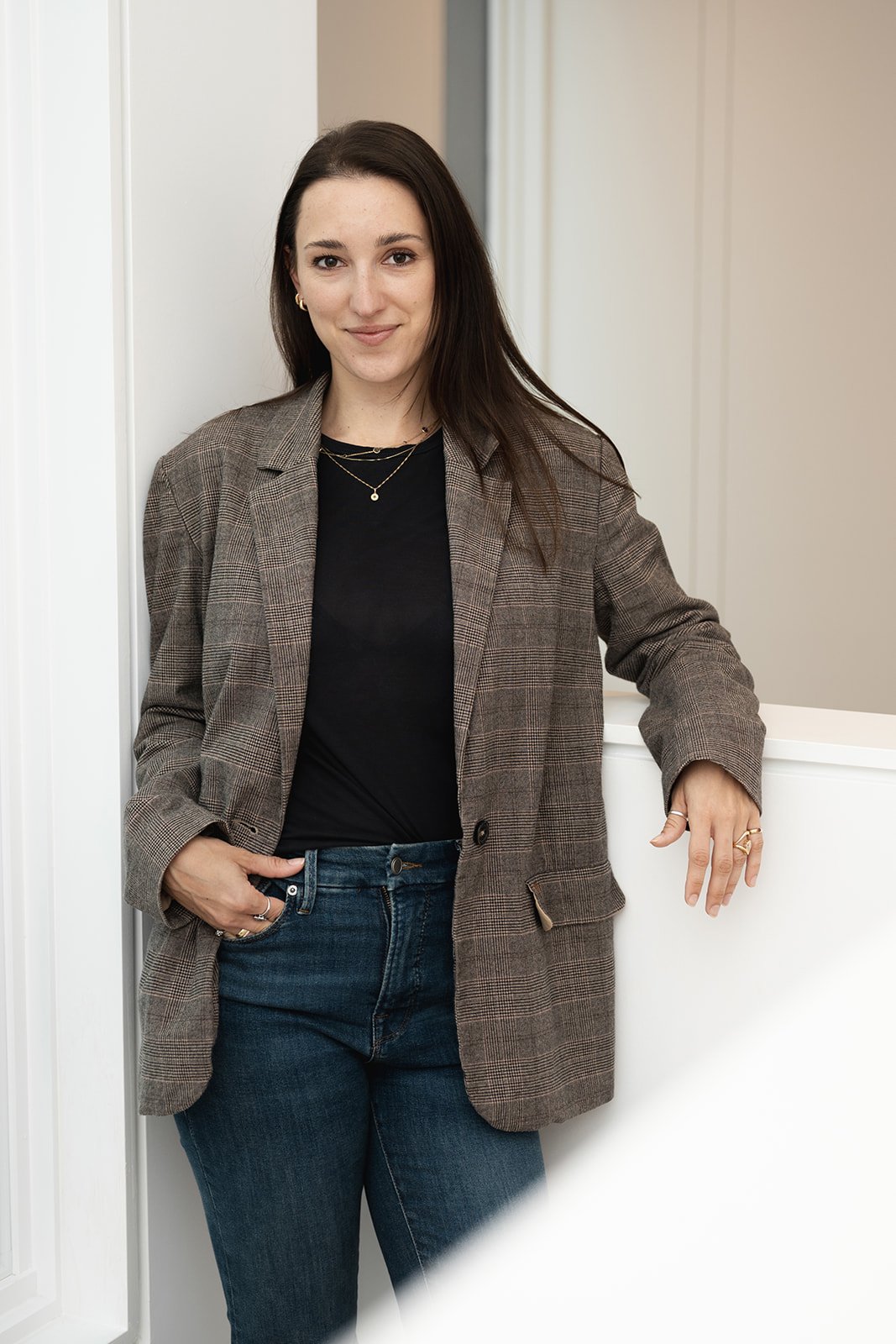 A woman with long dark hair, wearing a black top and a plaid blazer, stands against a white wall in a professional setting.