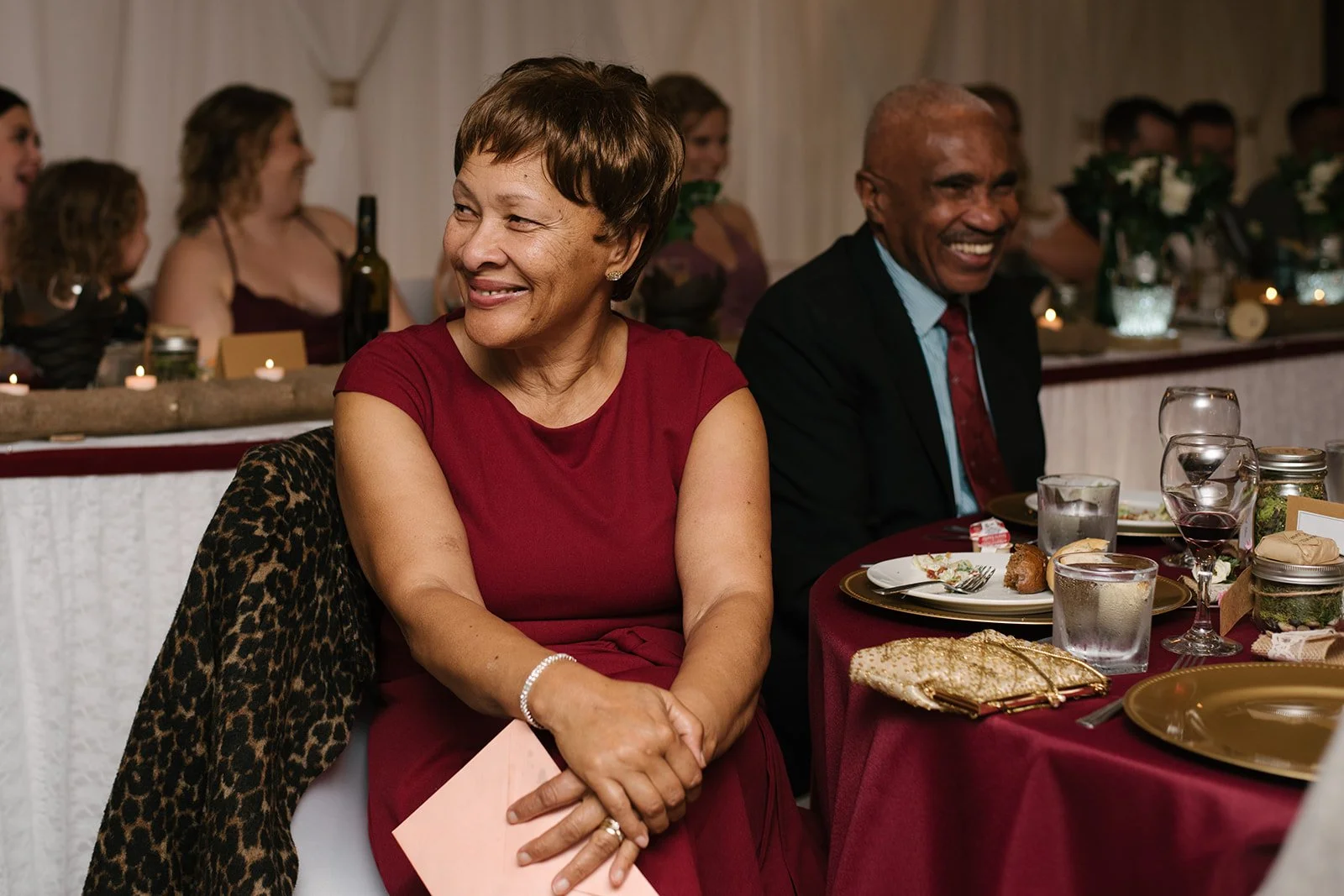A woman in a red dress smiling and sitting at a table with a man in a suit, during a wedding reception.