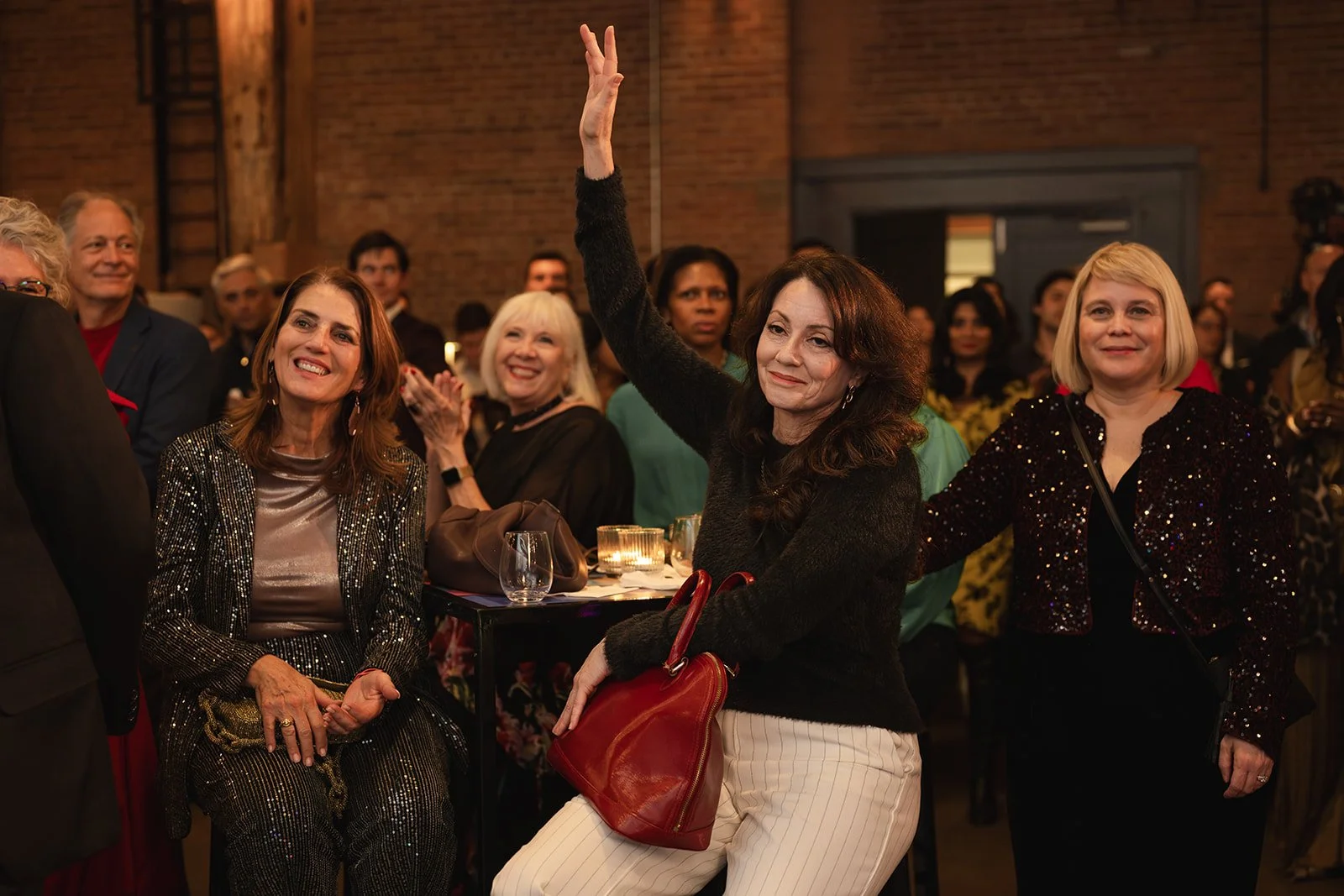 A group of women at a social event, with some smiling and one woman raising her hand. They are dressed in evening attire, surrounded by others in a warmly lit indoor setting with brick walls.